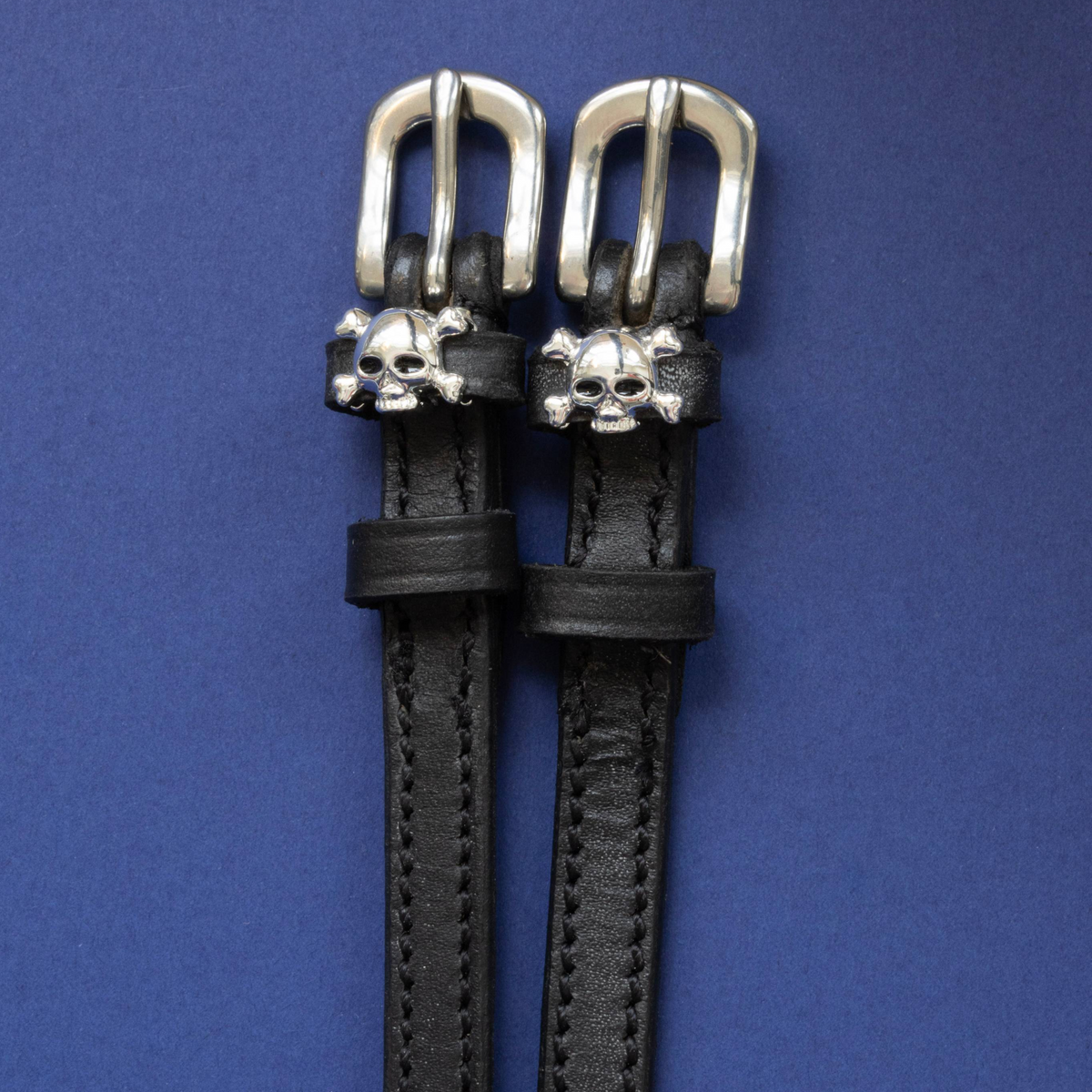 Black leather ManeJane spur straps with silver skull and crossbones charms on navy blue