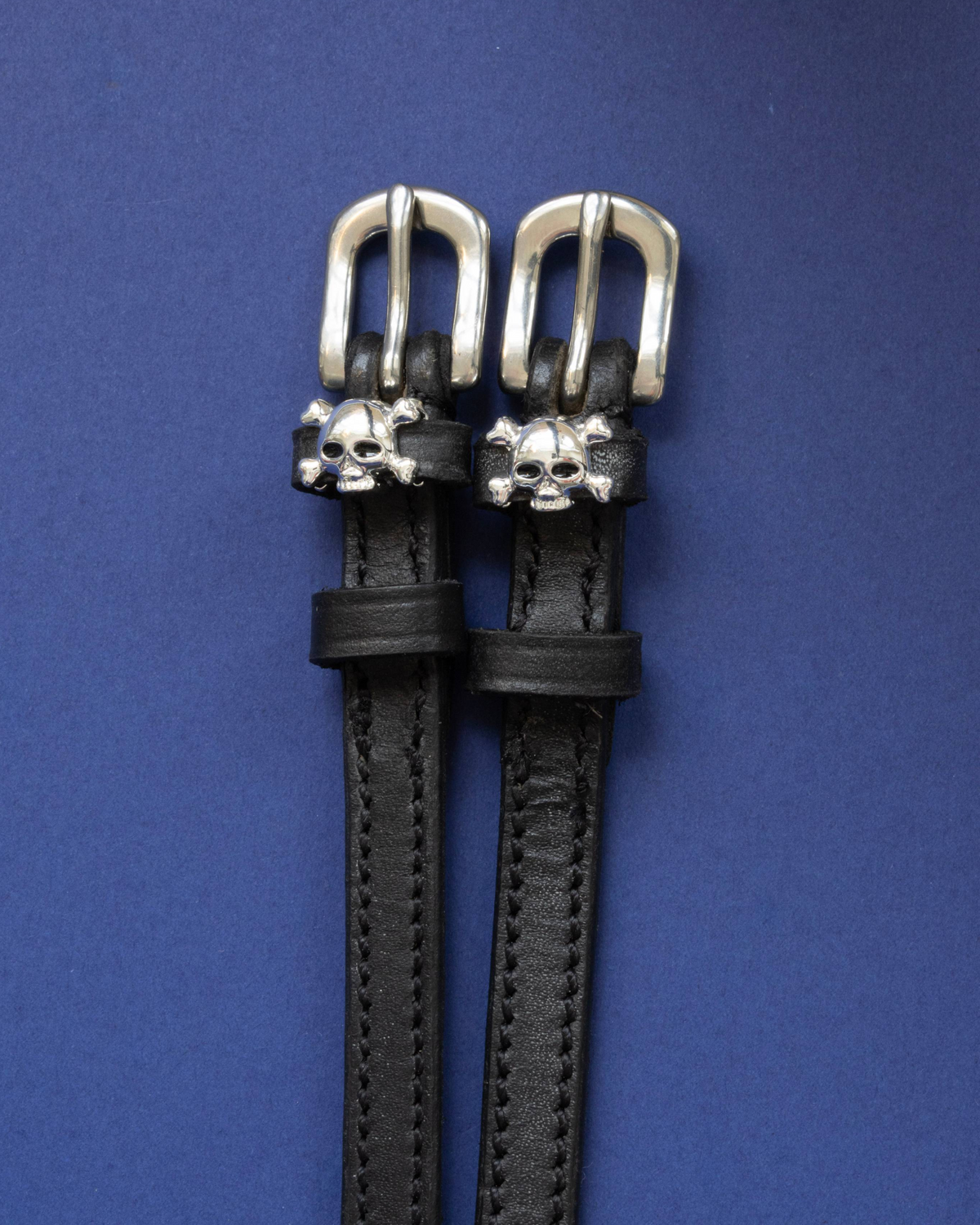 Black leather ManeJane spur straps with silver skull and crossbones charms on navy blue