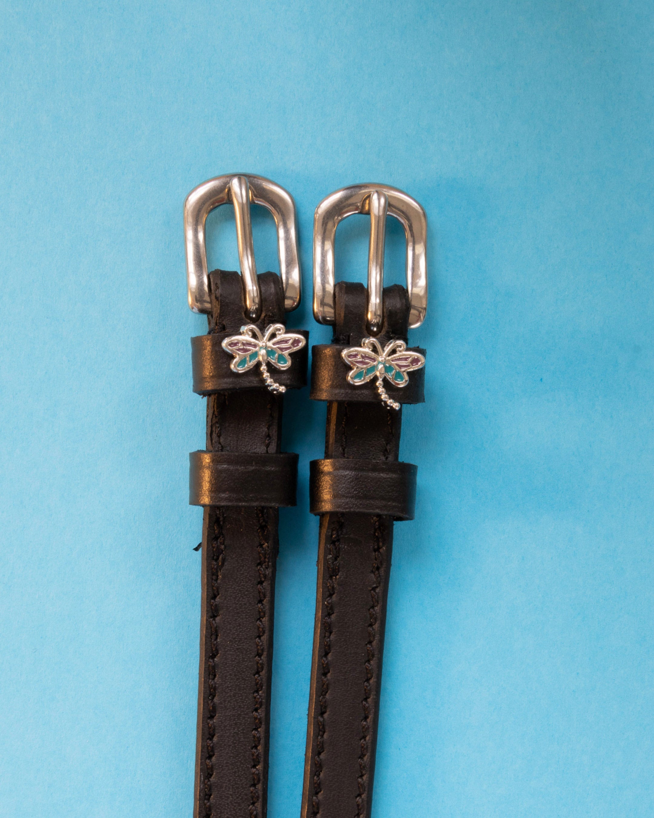 Dragonfly ManeJane spur straps with the signature Dragonfly charm, shown on a light blue background, by Dreamers & Schemers