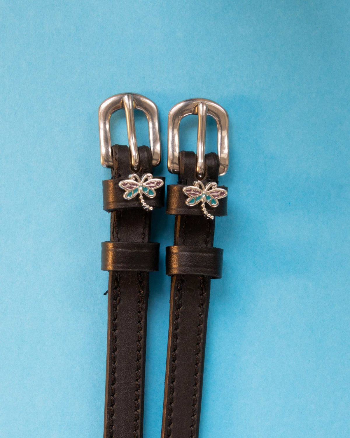 Dragonfly ManeJane spur straps with the signature Dragonfly charm, shown on a light blue background, by Dreamers & Schemers