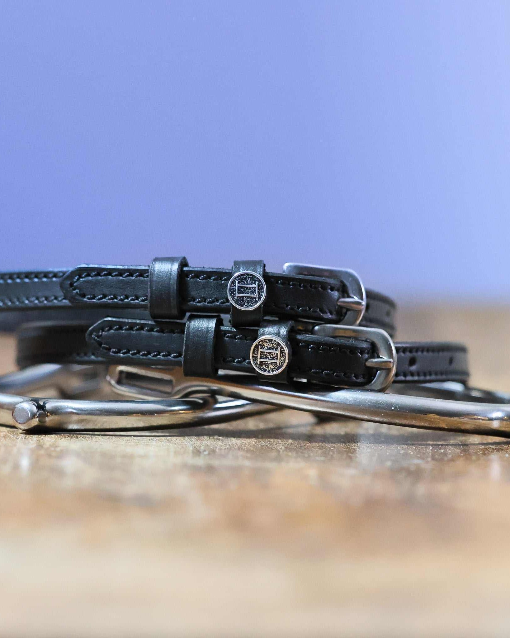 Black leather Gemini Spur Straps with stainless steel buckles and custom charms on wooden surface