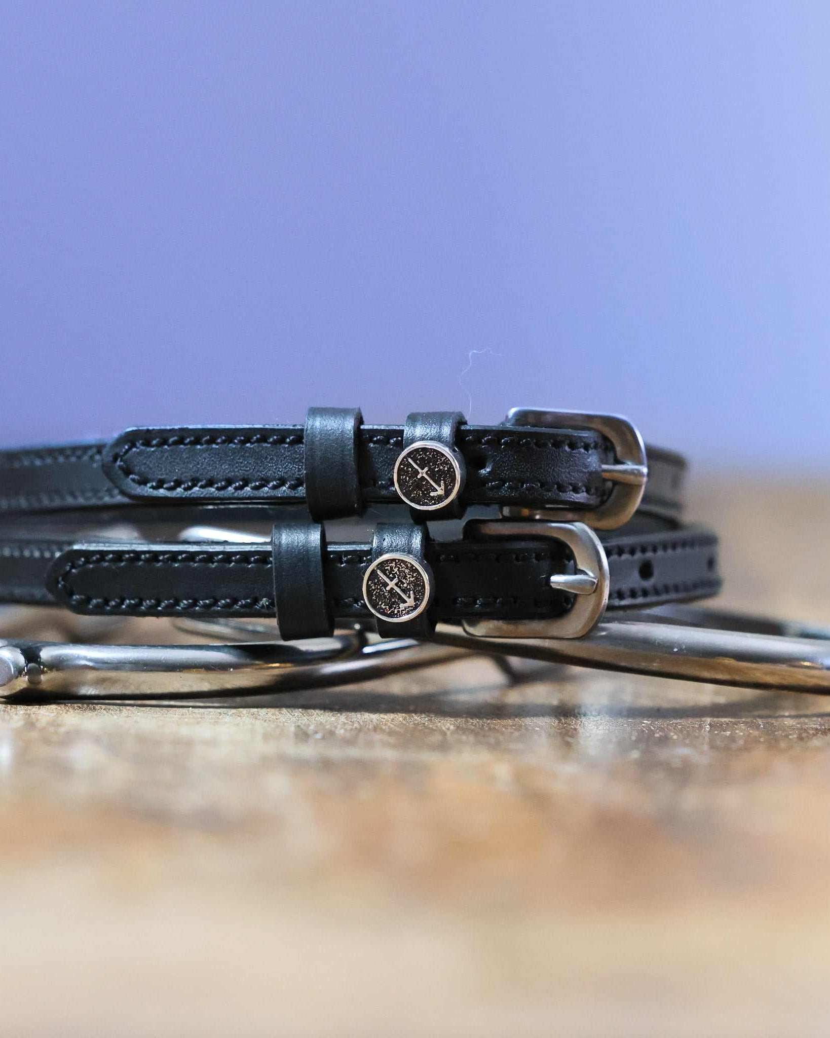 Black leather Sagittarius Spur Straps with stainless steel buckles and custom charm on wooden surface
