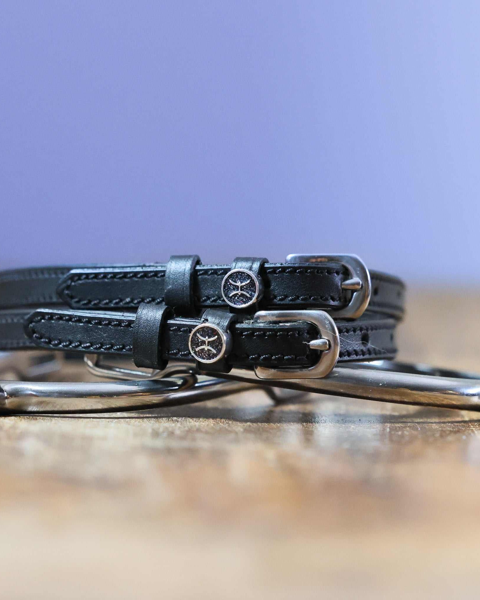 Black leather Pisces Spur Straps with stainless steel buckles and custom charm on wooden surface