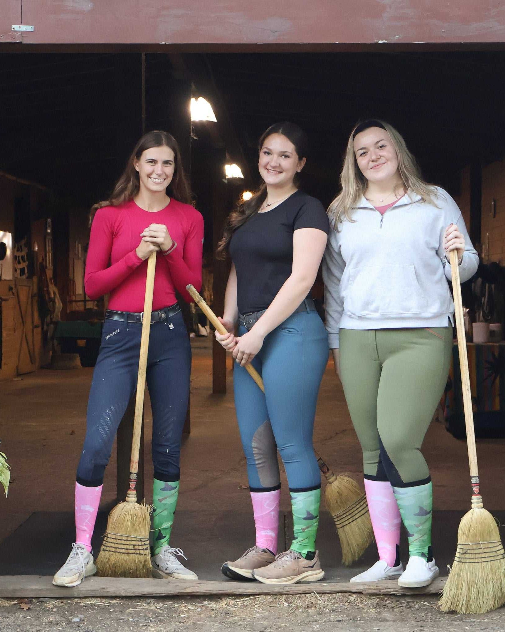 Three women wearing colorful boot socks standing with brooms in a barn entrance