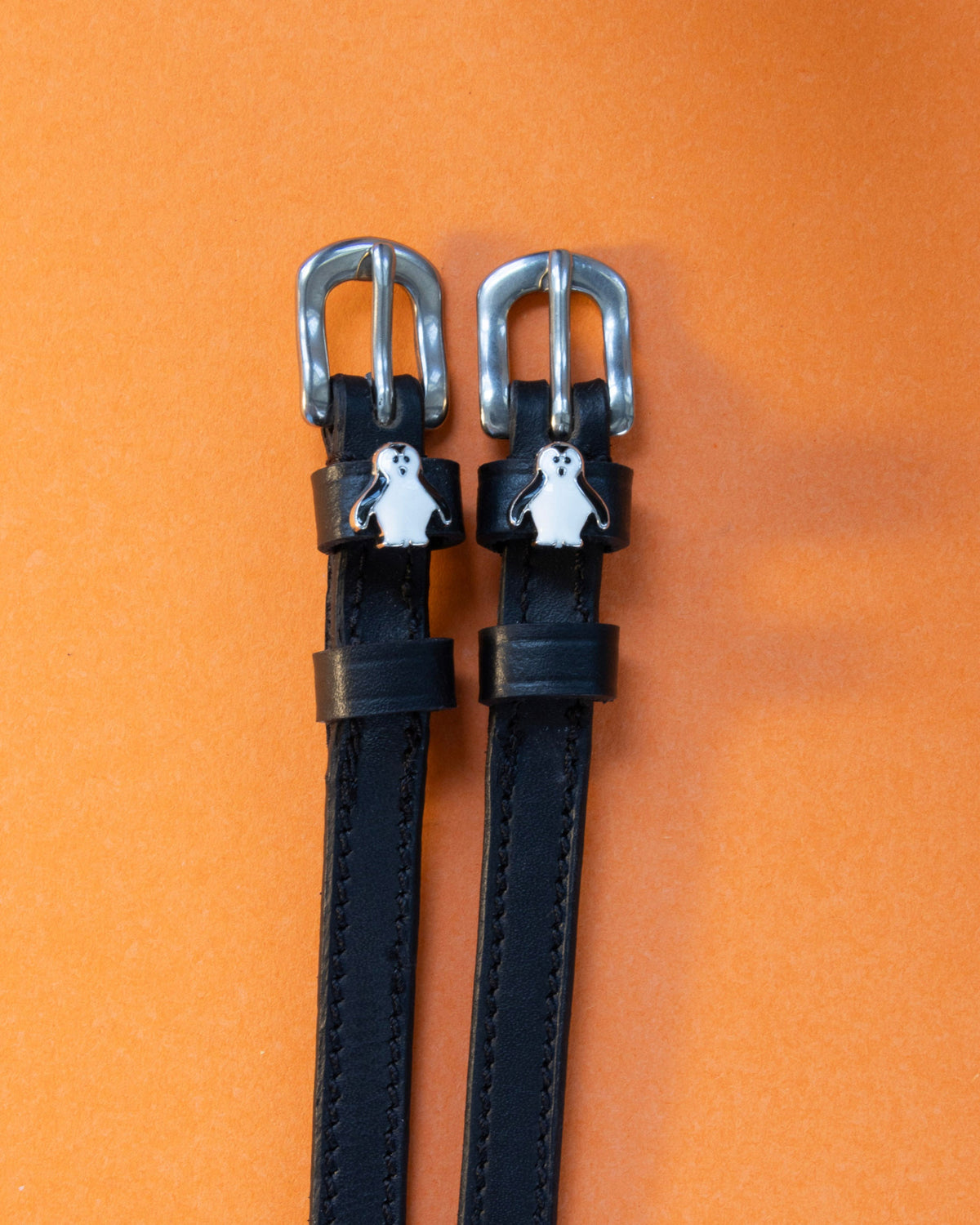 Penguin ManeJane spur straps with the signature Penguin charm, shown on an orange background