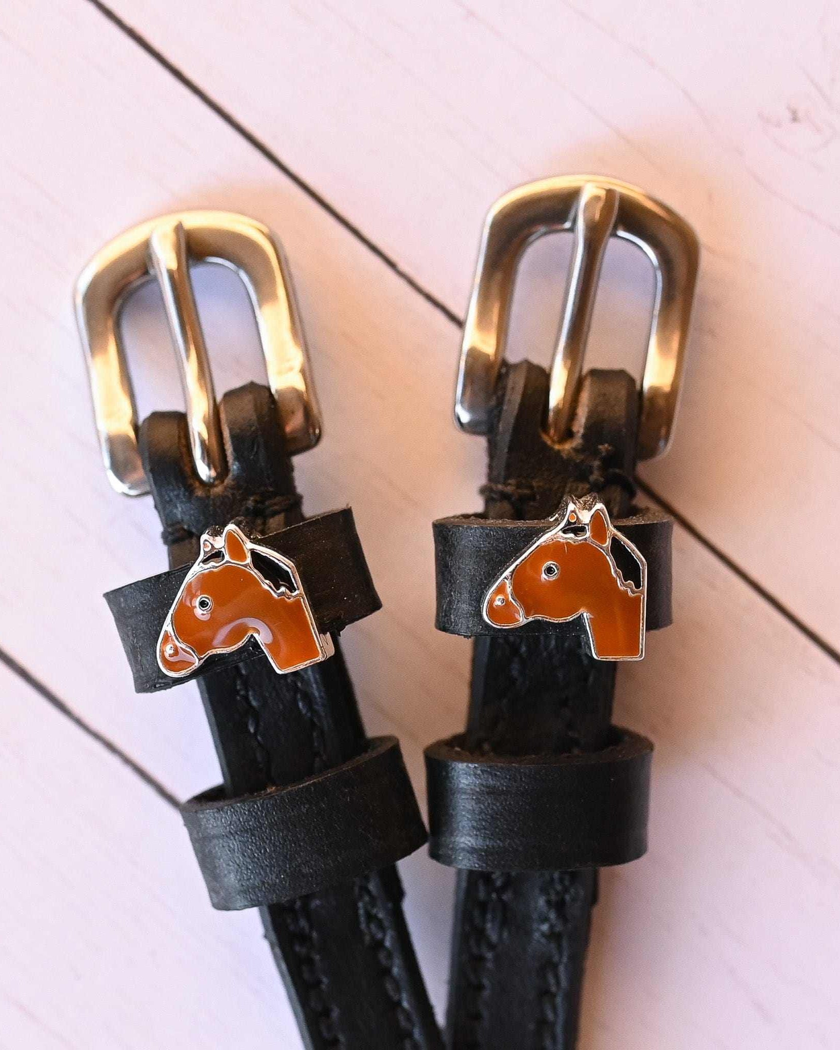 Close-up of tan horse-head charms and silver buckles on black spur straps, pink planks