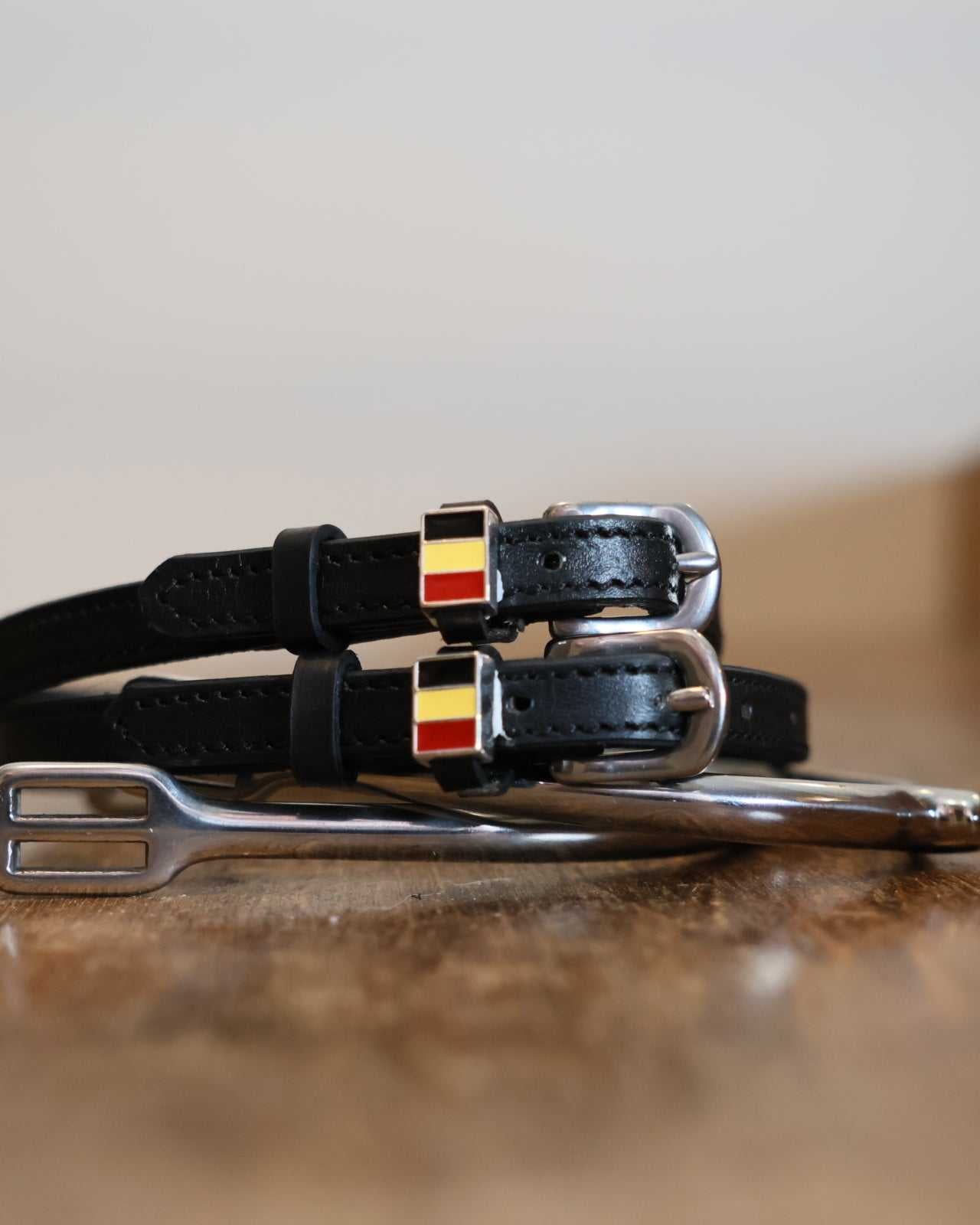 Black leather Belgium flag spur straps stacked on a silver spur on wood