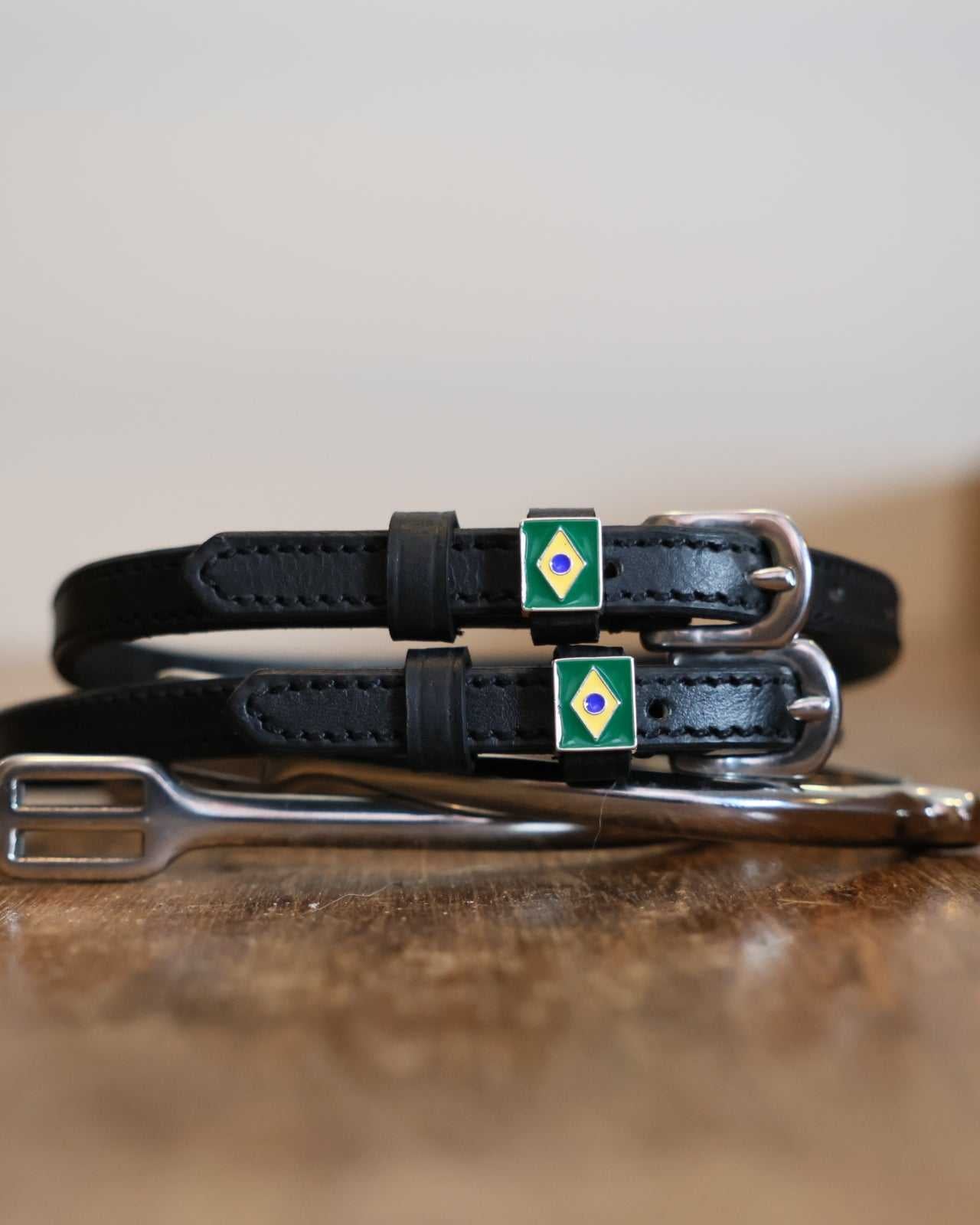 Black leather Brazil flag spur straps coiled beside a stirrup on a wooden surface
