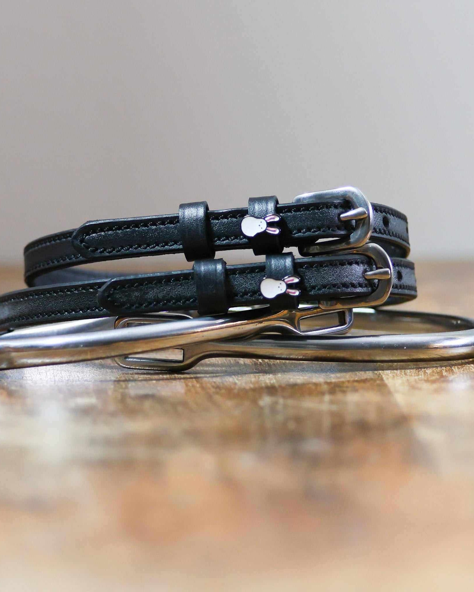 Stacked black Bunny Hop spur straps with silver buckles on wood beside a stirrup iron