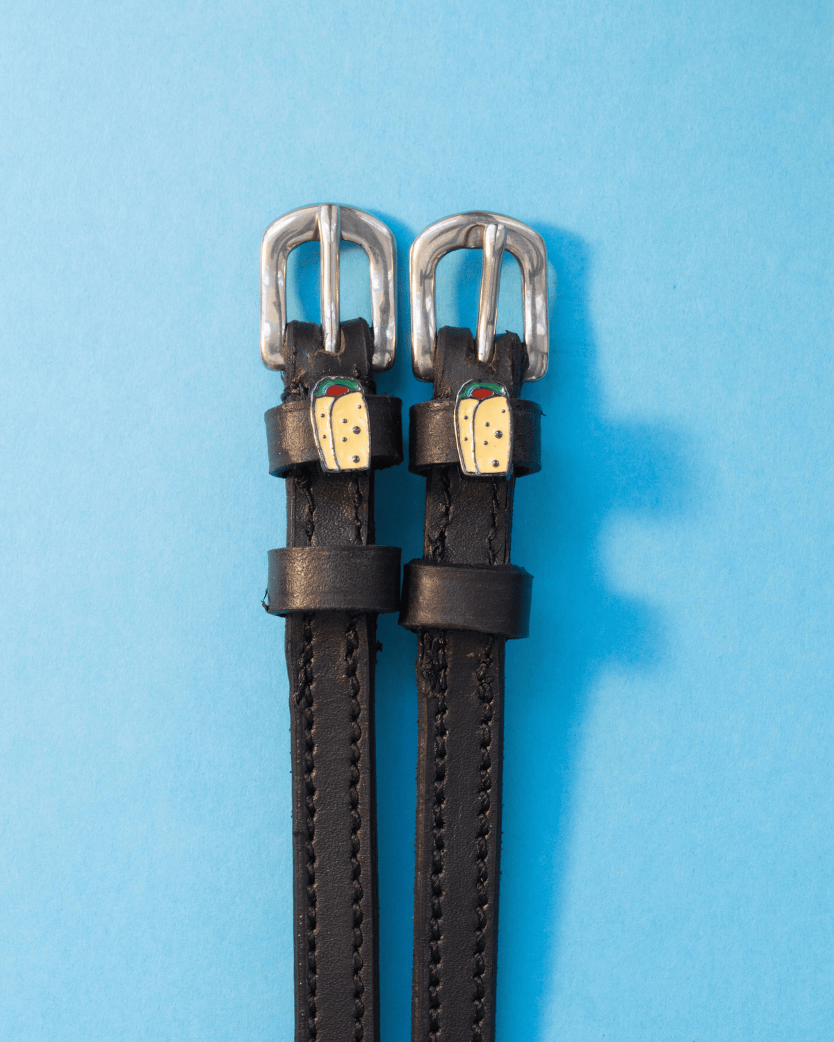 Pair of black leather spur straps with burrito charms flat-laid on a light blue background