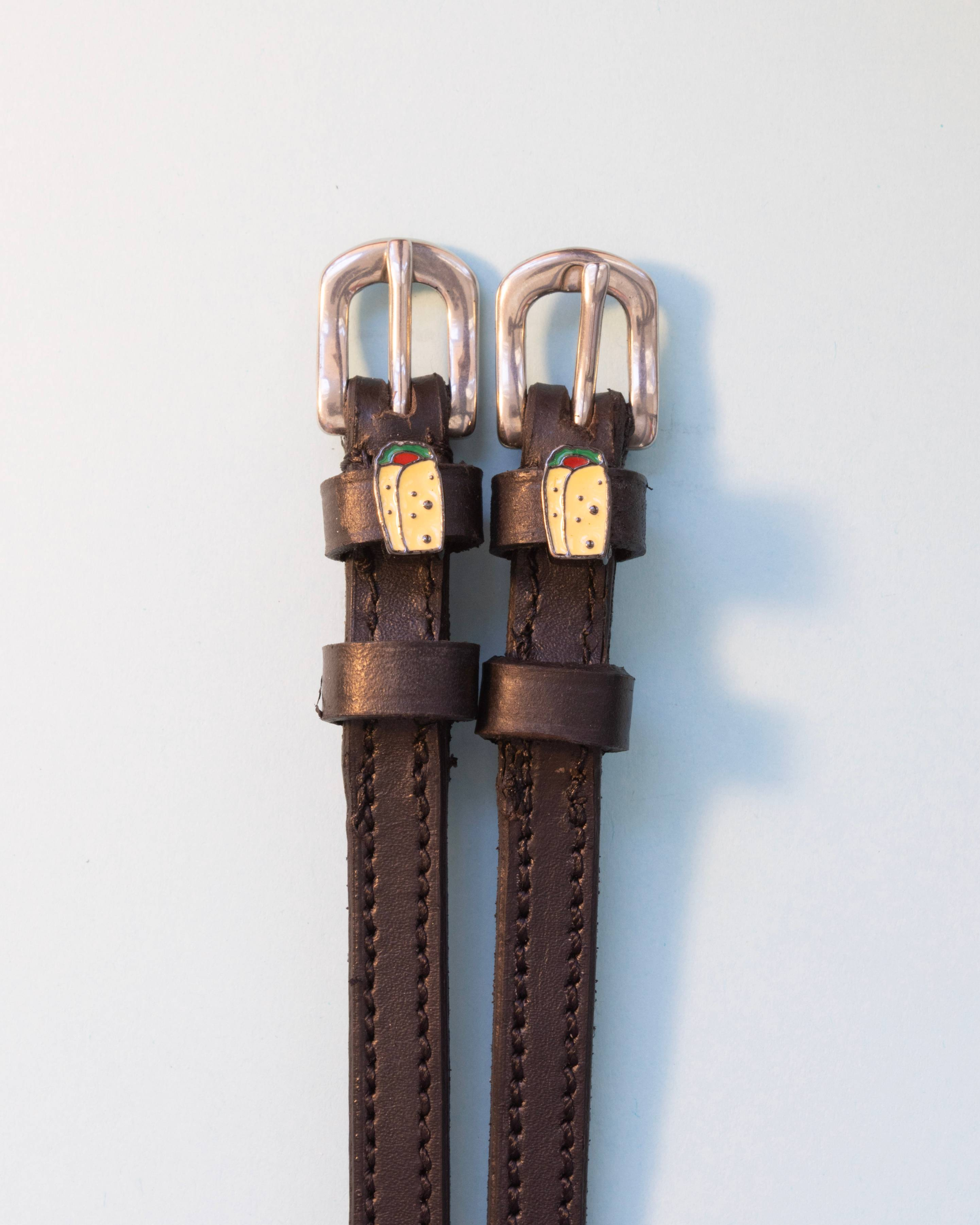 Brown leather ManeJane spur straps with silver buckles and burrito charms on white