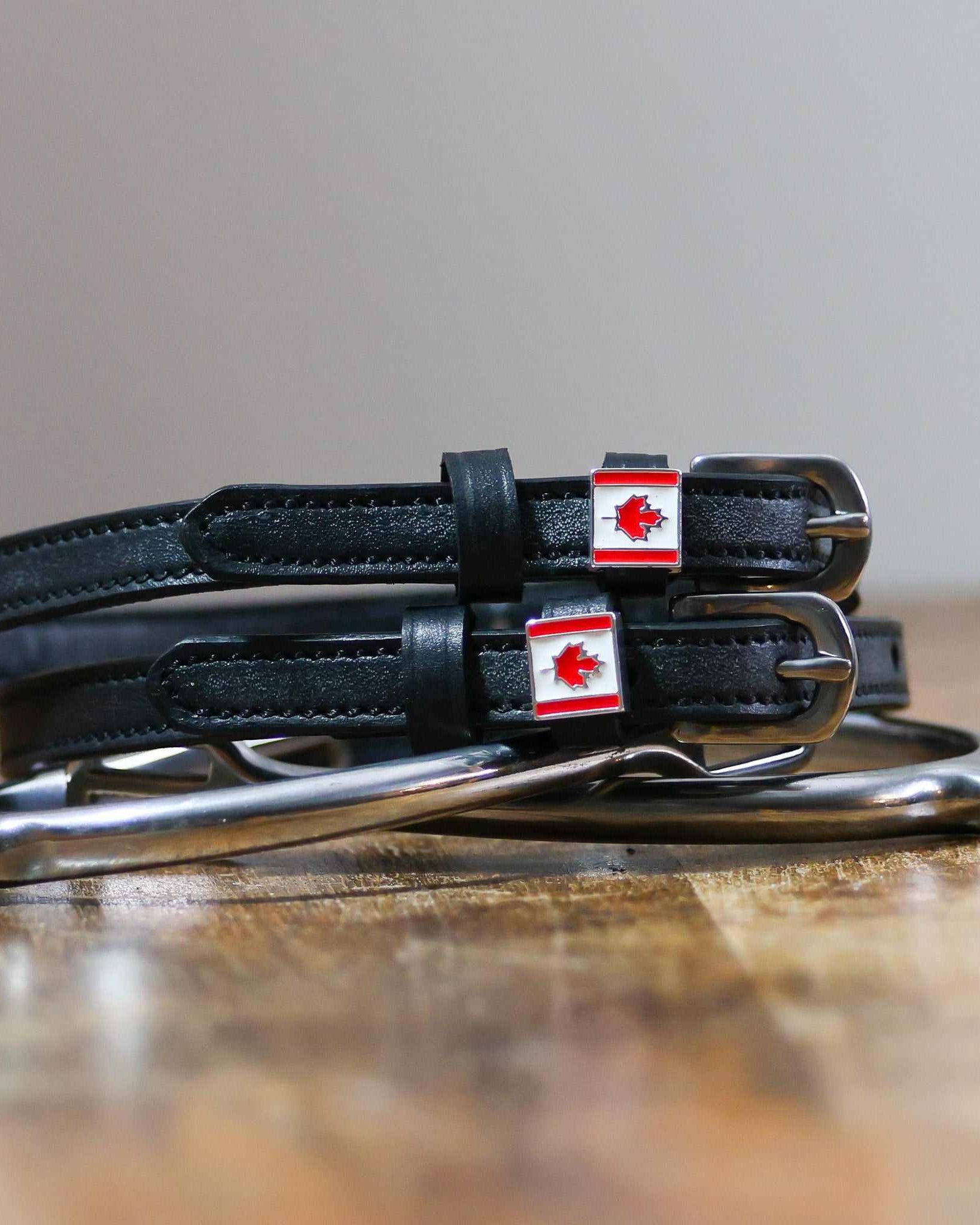 Black leather Canadian flag spur straps coiled beside a stirrup on a wooden surface