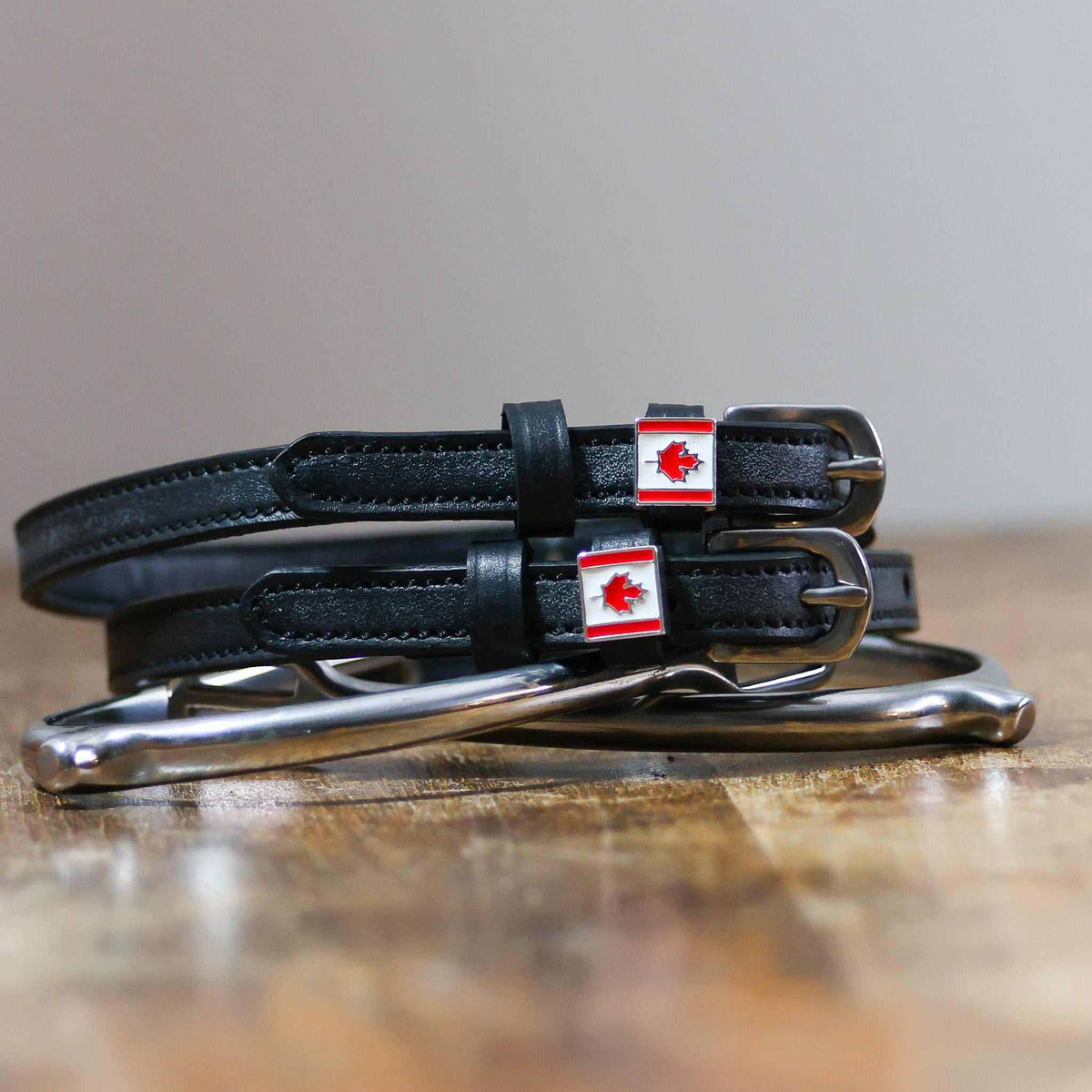 Black leather Canadian flag spur straps coiled beside a stirrup on a wooden surface