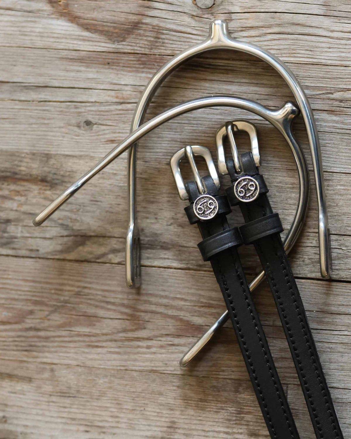 Overhead of black leather Cancer zodiac spur straps beside a silver spur on weathered wood