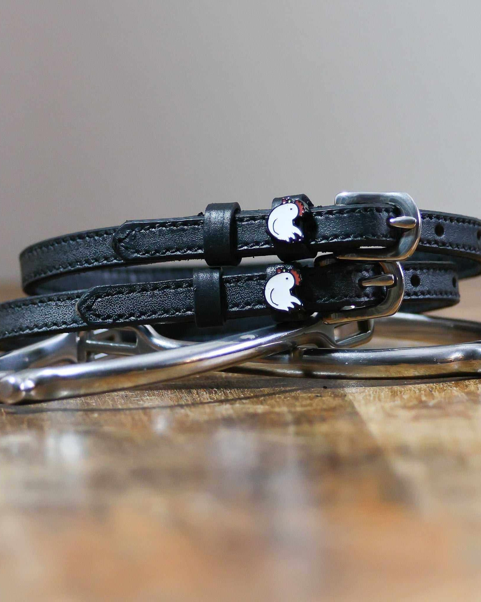 Black leather chicken spur straps coiled beside a silver stirrup on a wooden surface