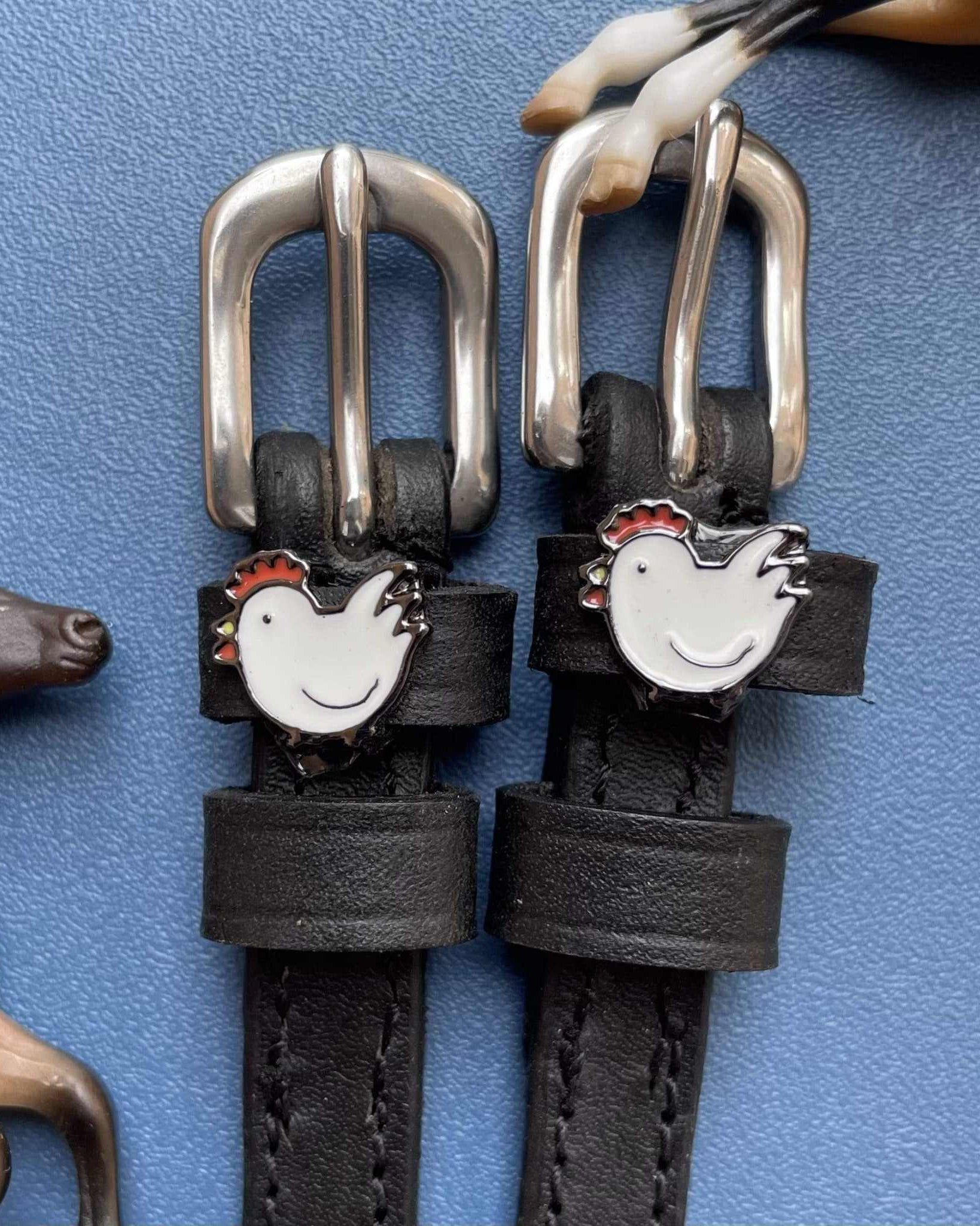 White hen charm spur straps with silver buckles beside a horse figurine on blue