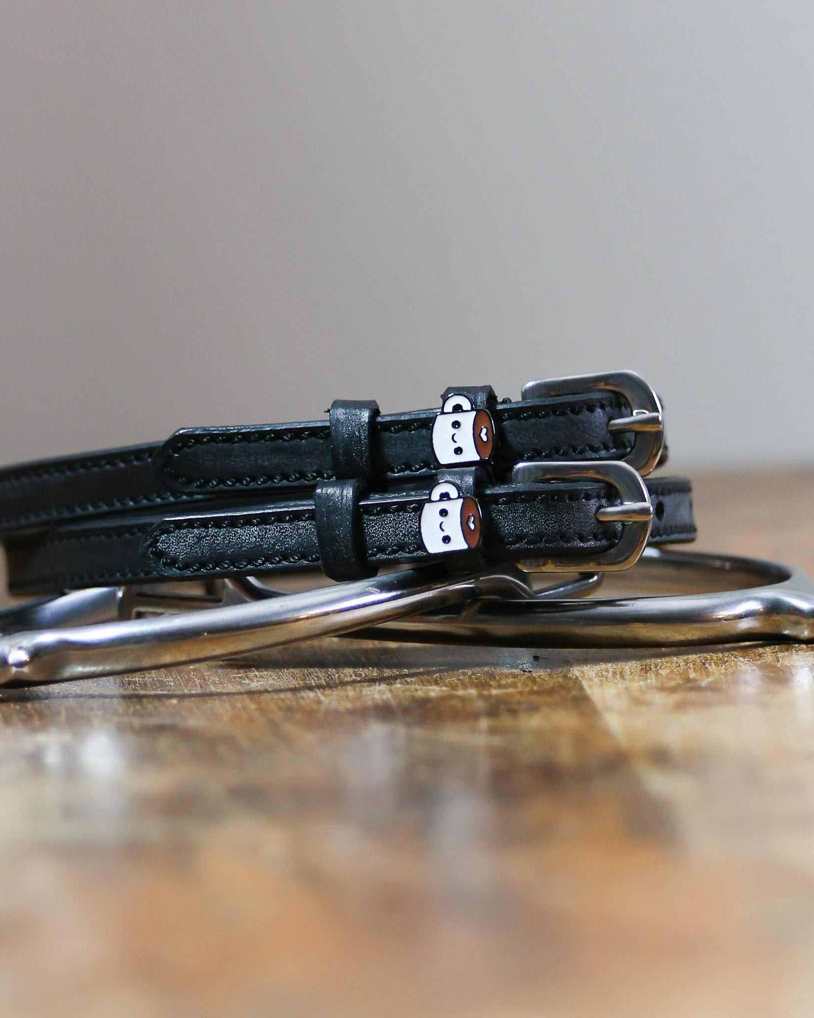 Black leather coffee-mug spur straps coiled on wood beside a stirrup