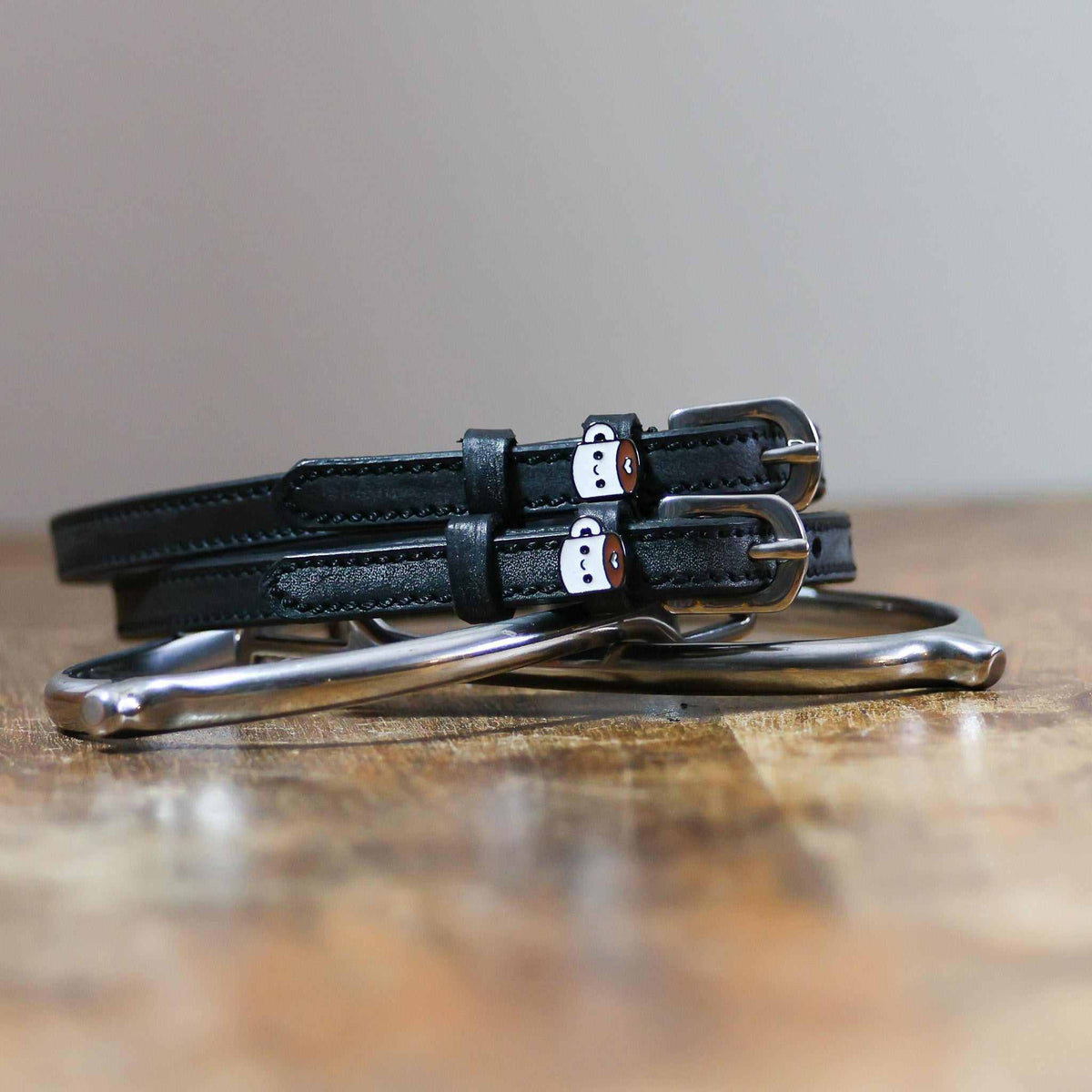 Black leather coffee-mug spur straps coiled on wood beside a stirrup