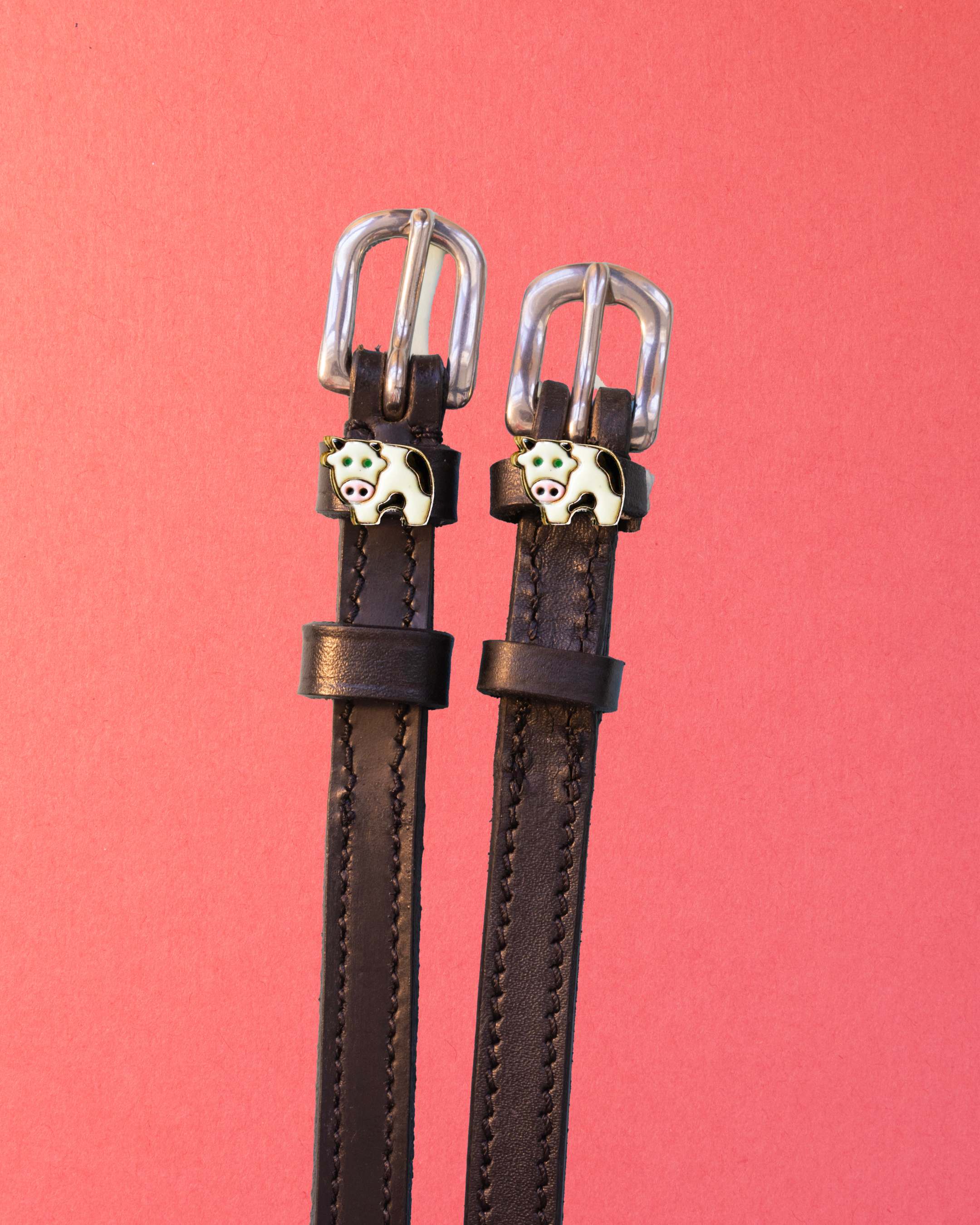 Pair of black leather ManeJane cow spur straps with enamel cow charms on a coral background