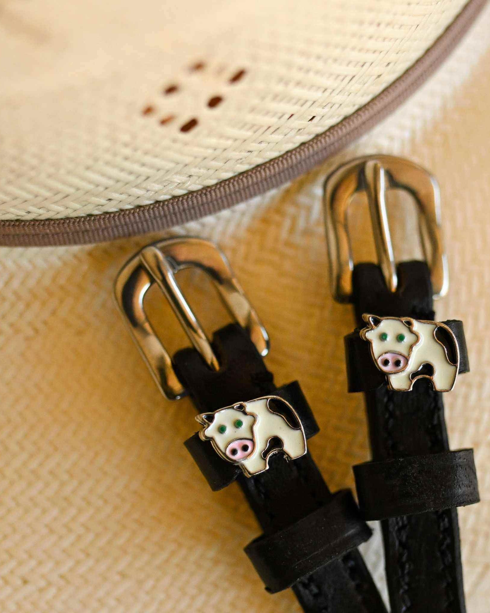 Black leather cow spur straps beside a straw sun hat, ManeJane enamel cow charm close-up