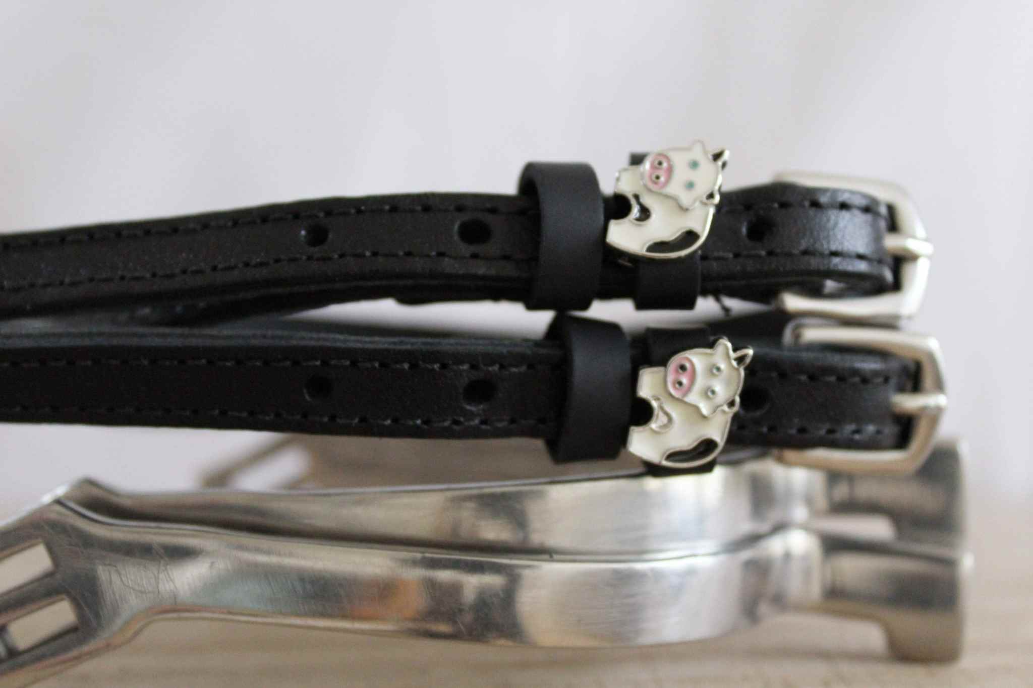 Side view of black leather ManeJane cow spur straps with cow charms resting on a silver stirrup iron