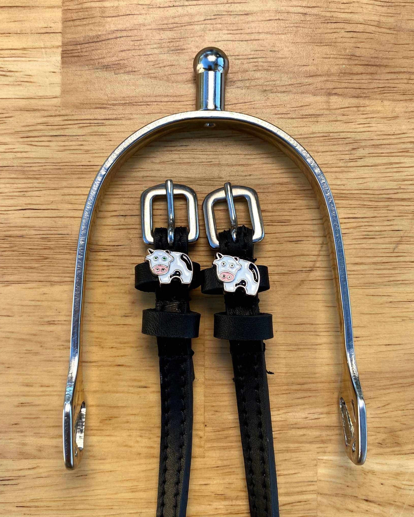 Black leather cow spur straps laid over a silver spur on a wooden surface, ManeJane cow charms