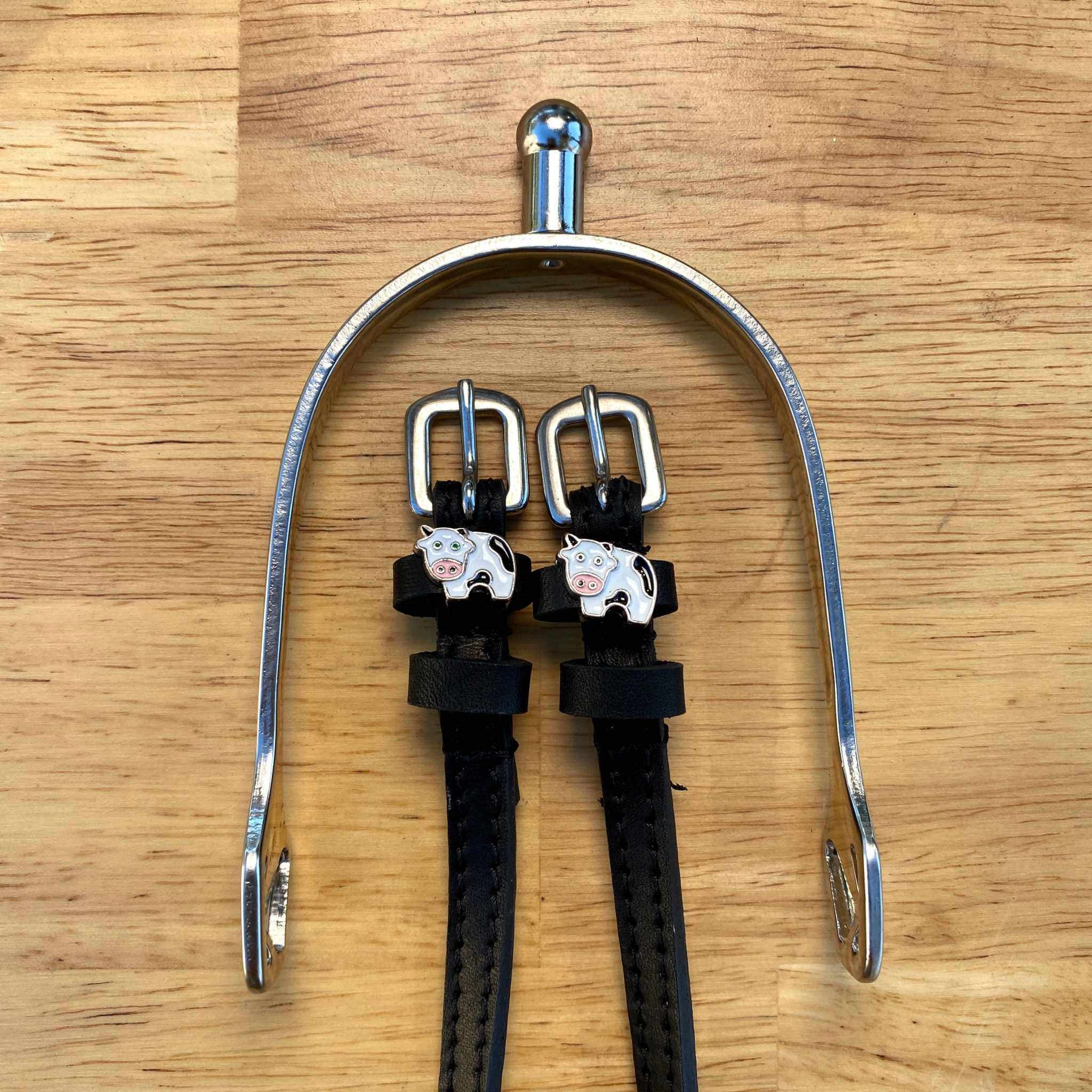 Black leather cow spur straps laid over a silver spur on a wooden surface, ManeJane cow charms