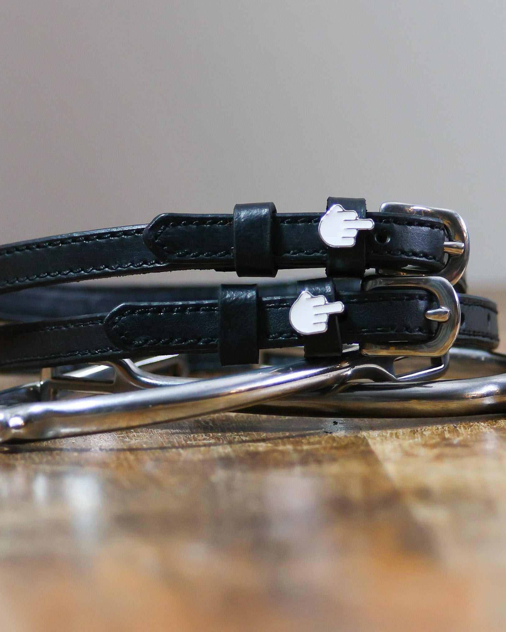 Stacked black Da Bird spur straps with silver buckles on wood beside a stirrup iron, ManeJane