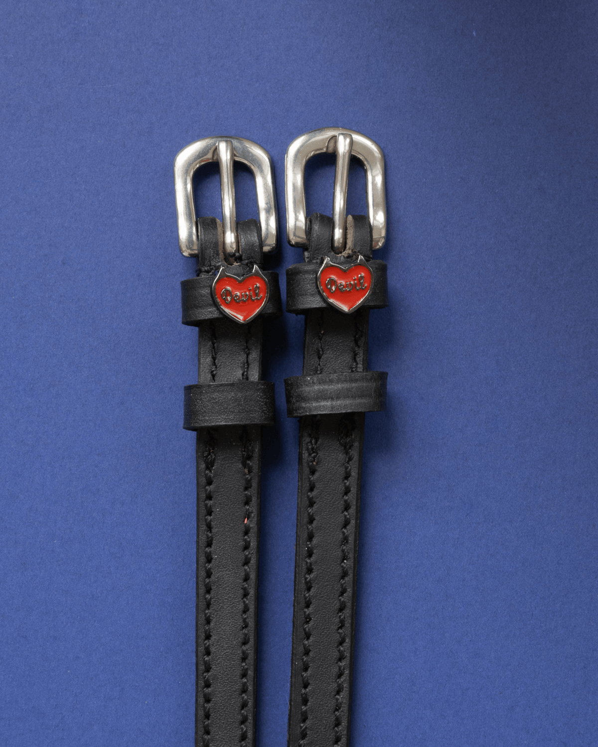 Pair of black leather Devil heart spur straps flat-laid on a royal blue background