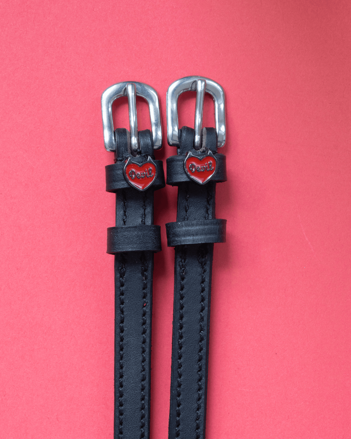Black leather Devil heart spur straps with silver buckles on a coral background
