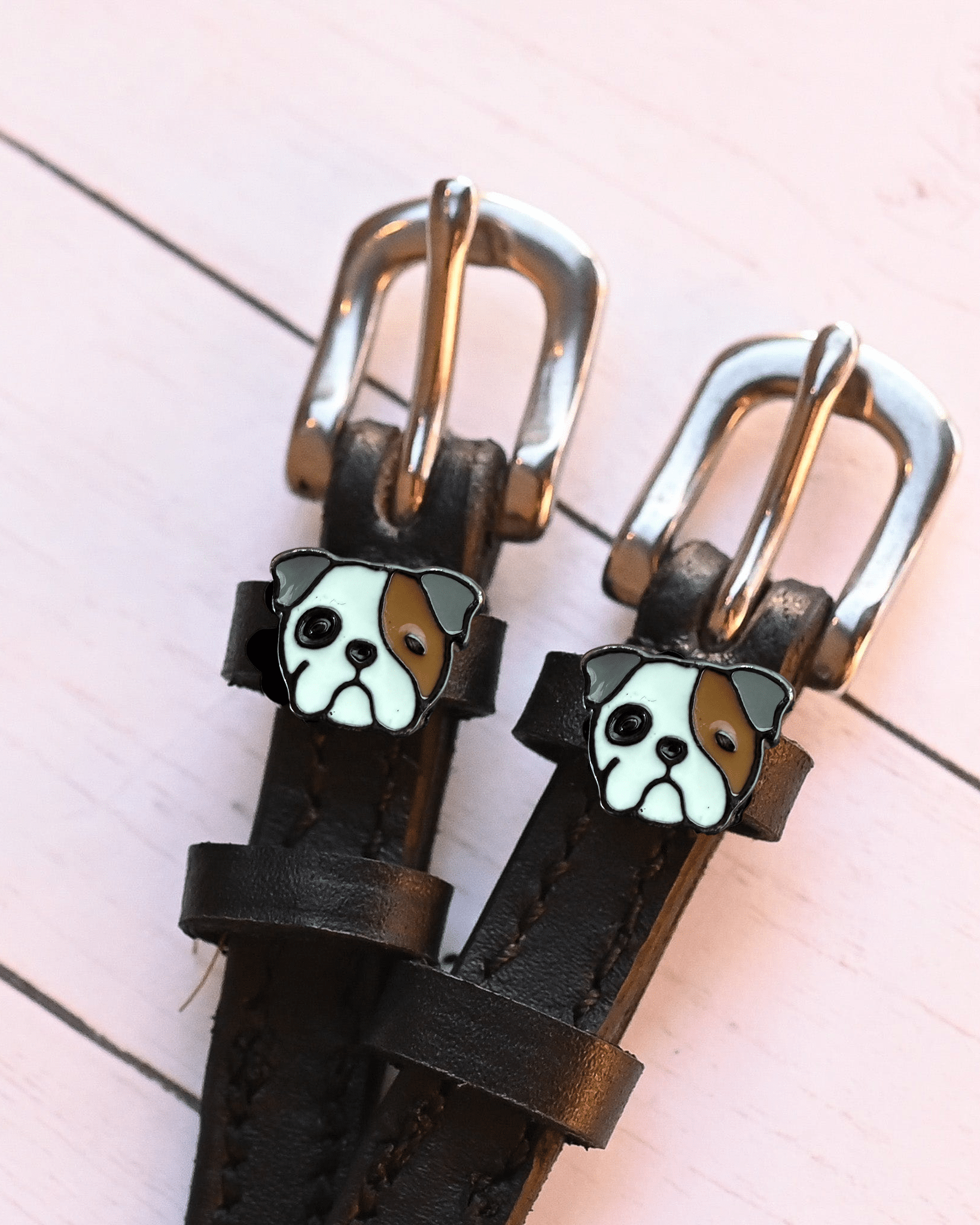 Bulldog head charms on dark brown leather spur straps over pale pink wood planks