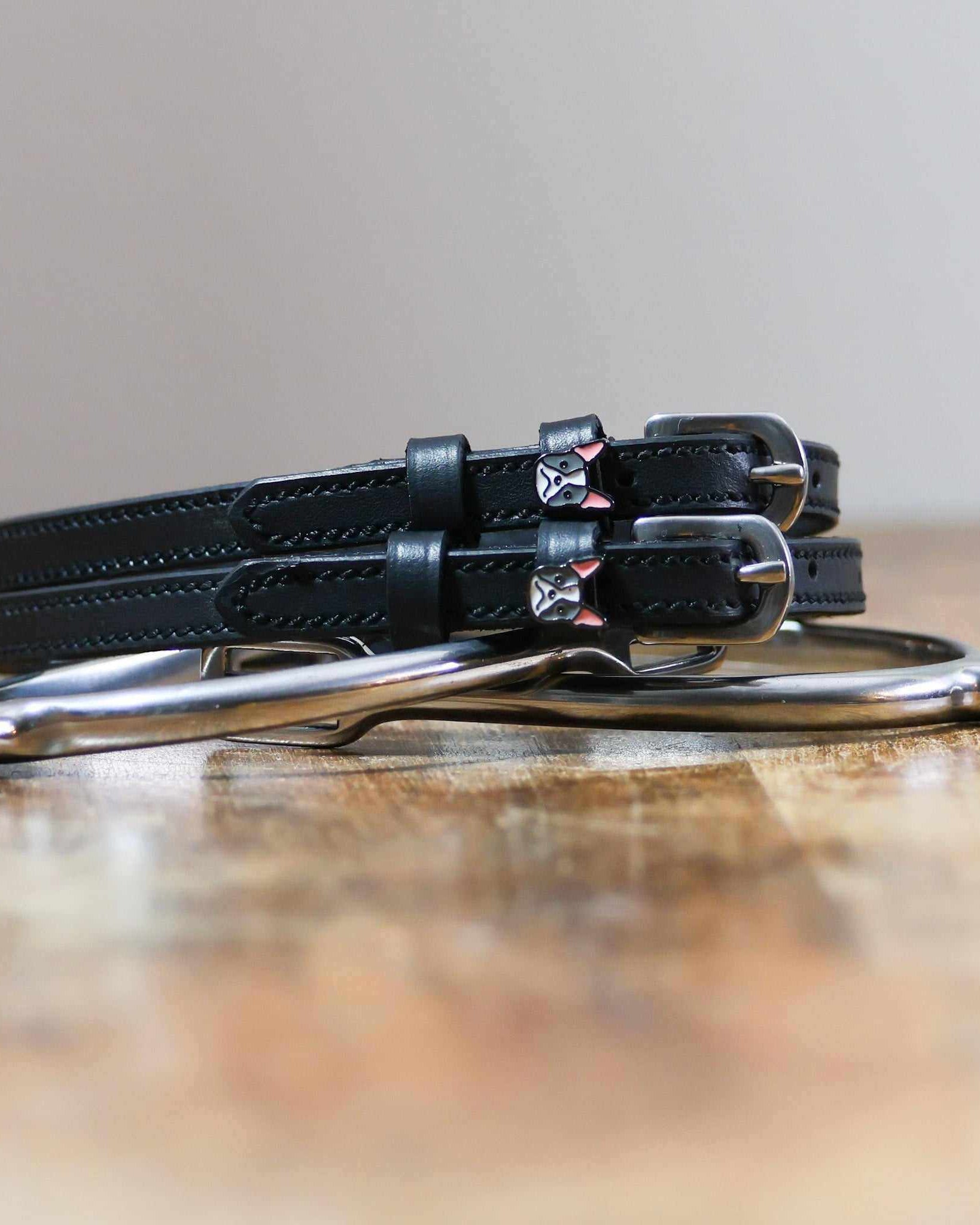 Black leather Frenchie spur straps coiled beside a stirrup on a wooden surface