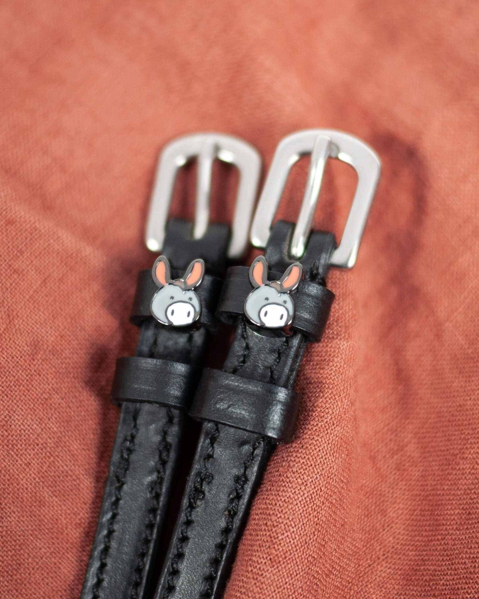 Black leather donkey spur straps with silver buckles angled on a rust linen background