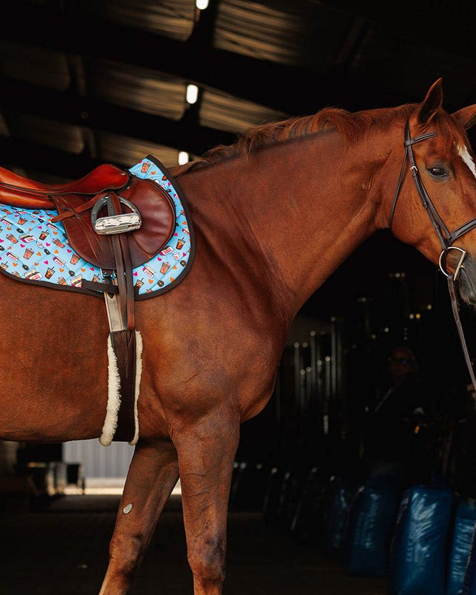 Chestnut horse wearing a blue donut-themed saddle pad and brown saddle in stable
