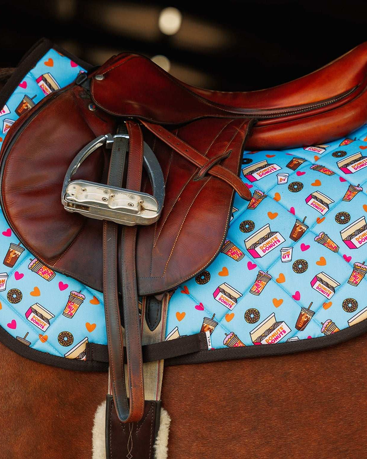 Colorful donut-themed saddle pad under brown leather horse saddle and stirrup