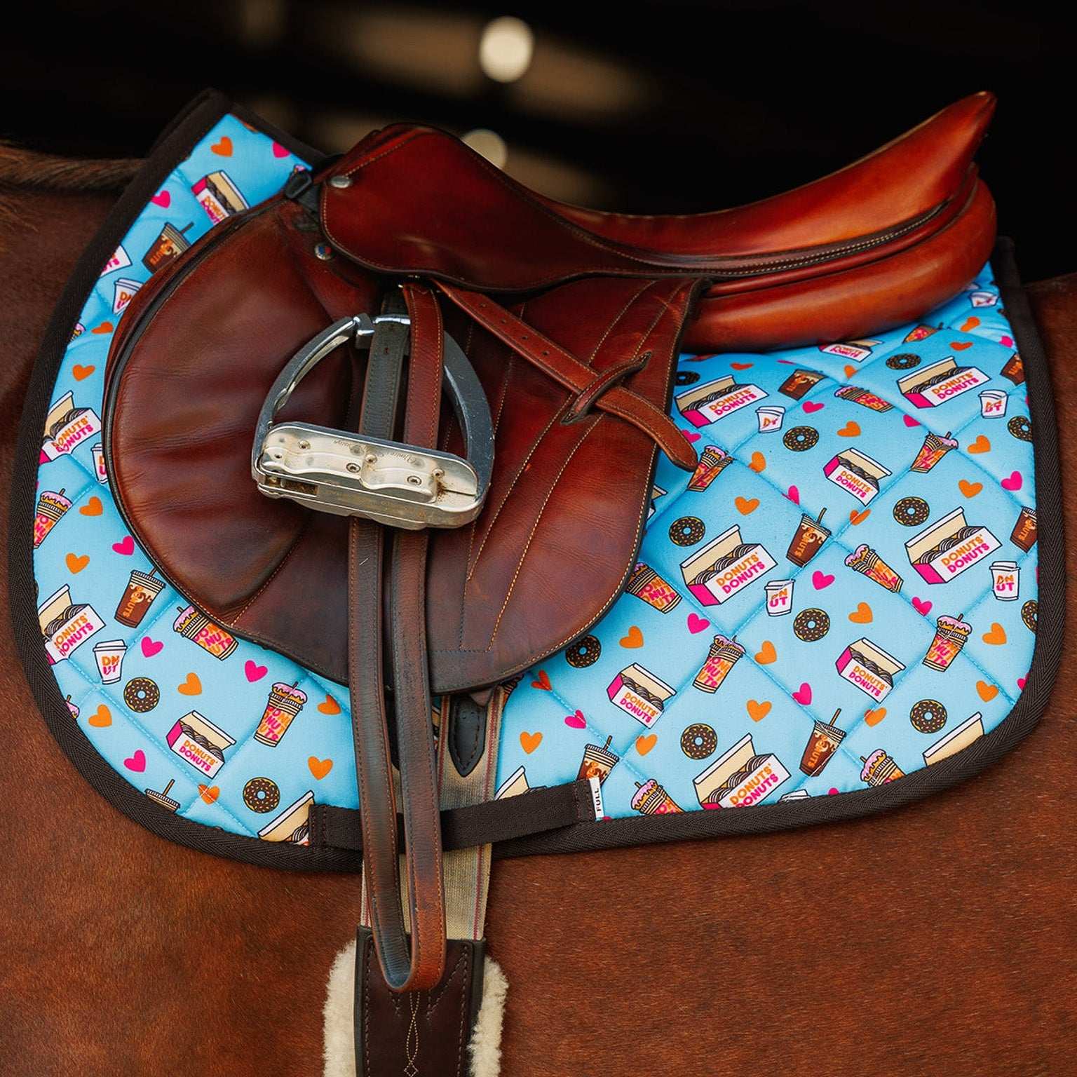Colorful donut-themed saddle pad under brown leather horse saddle and stirrup