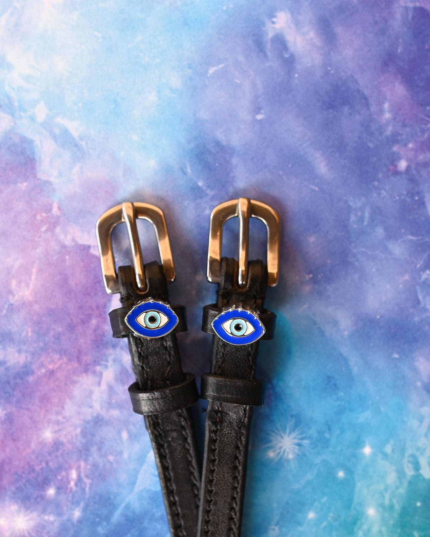 Black leather evil eye spur straps with brass buckles on a galaxy print background