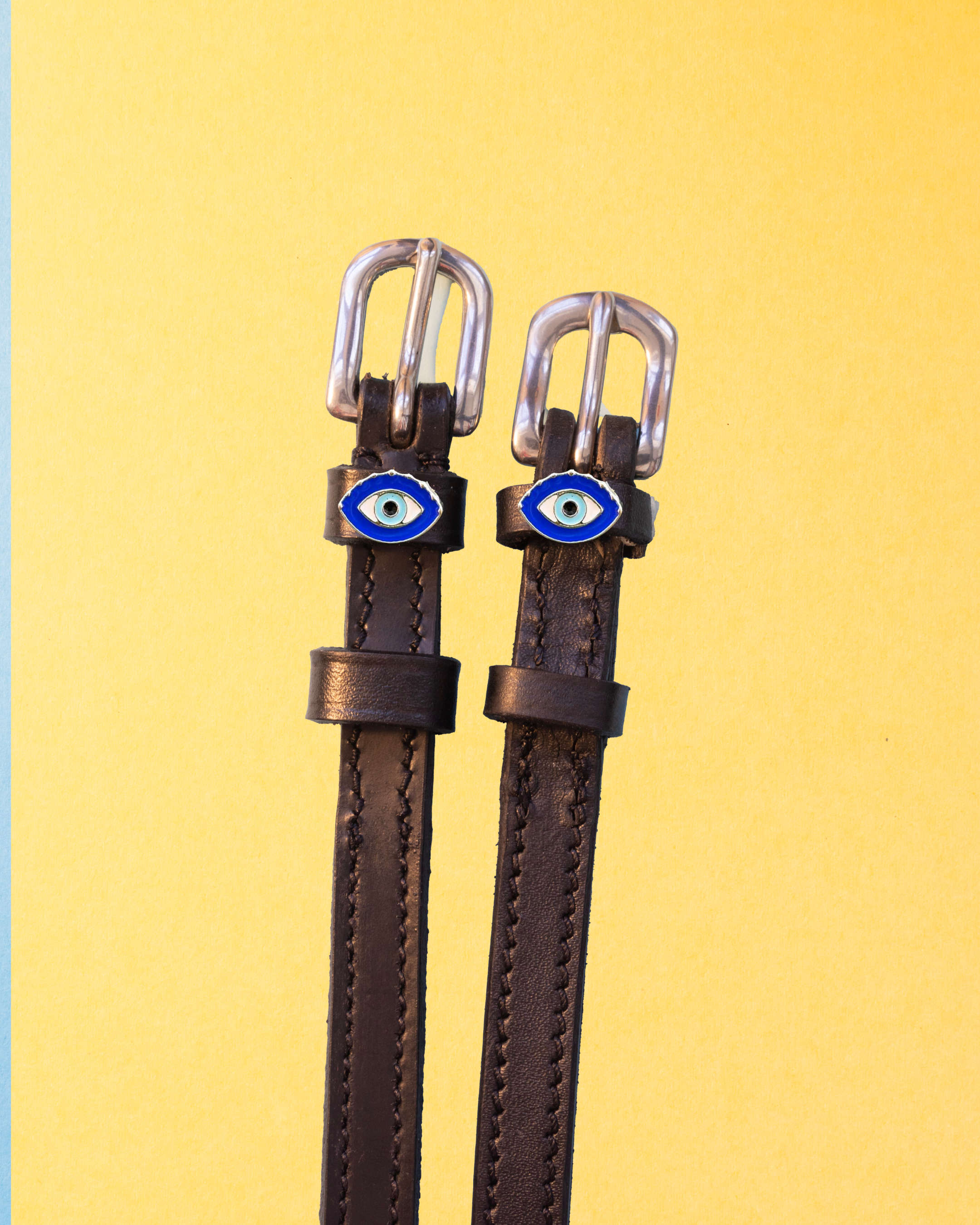 Brown leather ManeJane evil eye spur straps with silver buckles on a yellow background