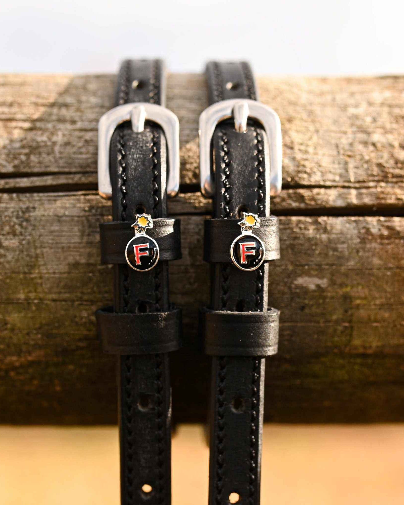 Black leather F Bomb spur straps hanging over a weathered wood rail with bomb charms