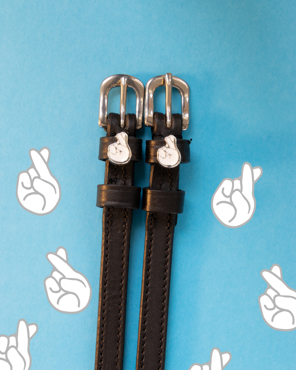 Black leather crossed-fingers spur straps flat-laid on a blue crossed-fingers icon backdrop