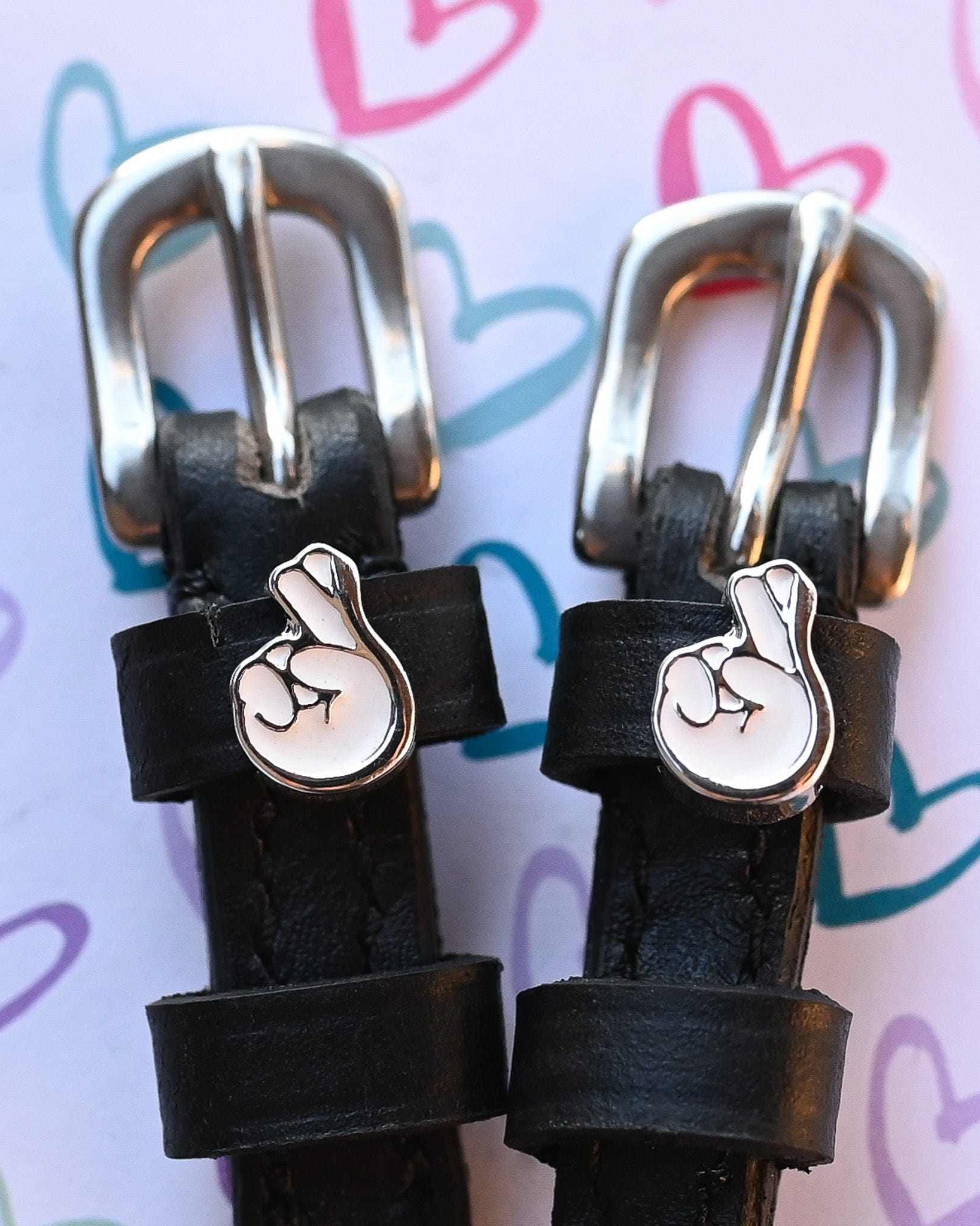 Silver crossed-fingers charms on black spur strap keepers against a hand-drawn hearts print