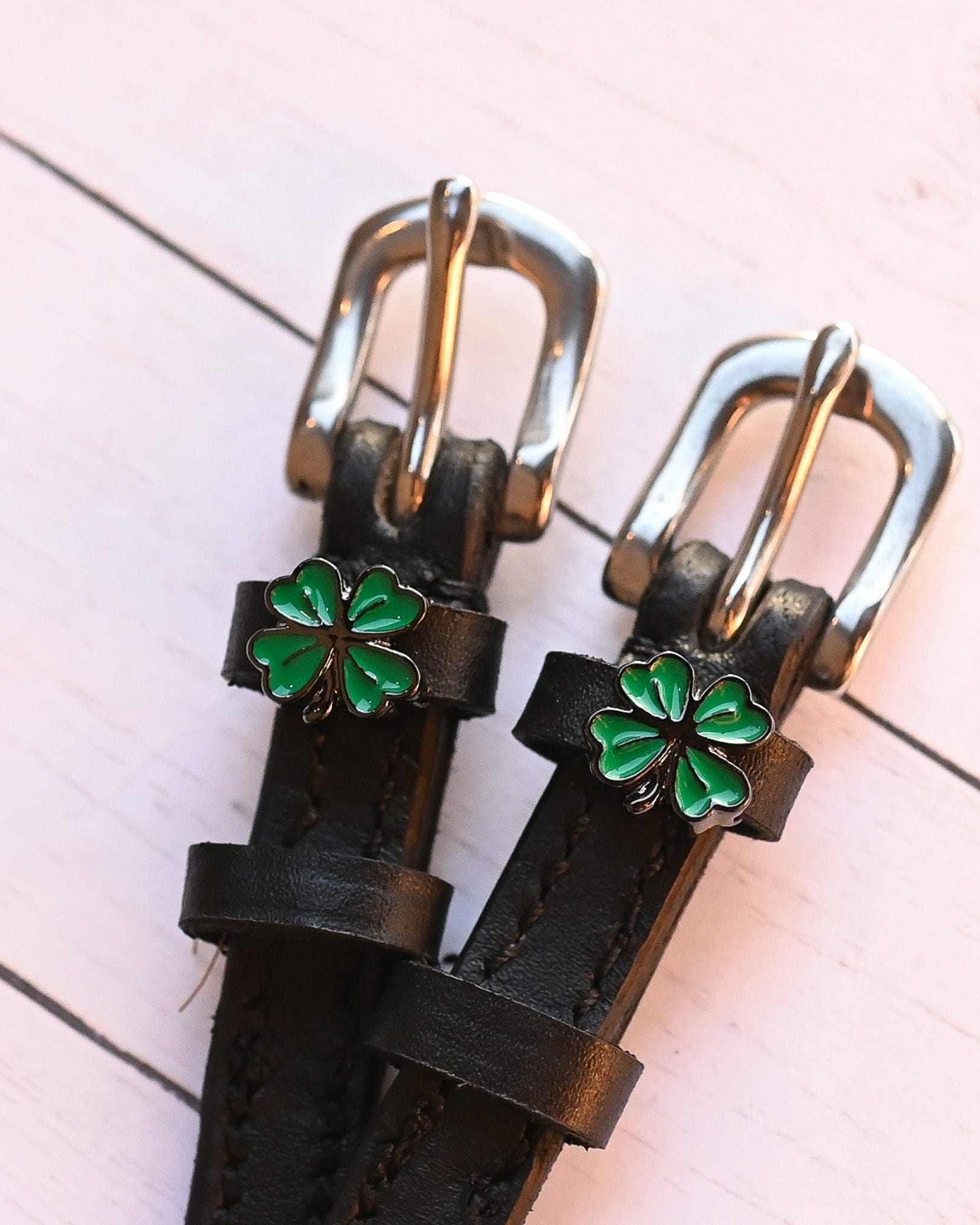 Angled close-up of ManeJane clover spur straps with green charms on pink wood planks