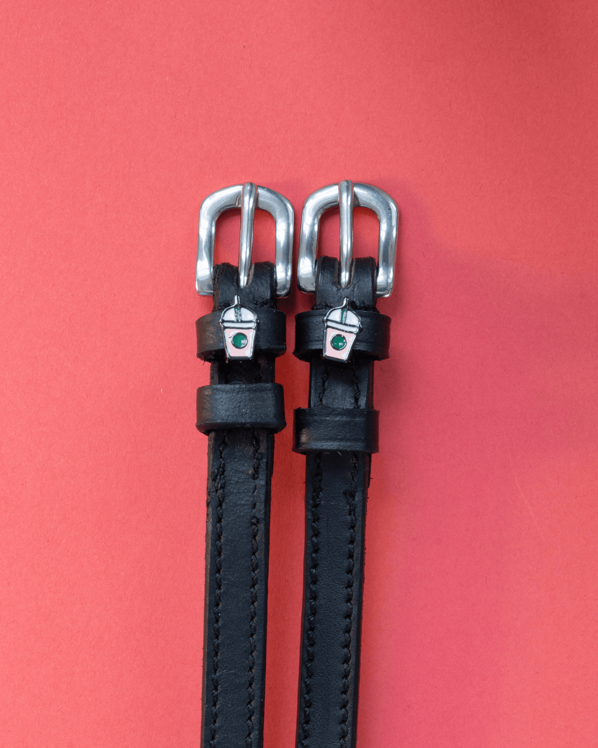 Black leather ManeJane spur straps with silver frappe cup charms on a red background