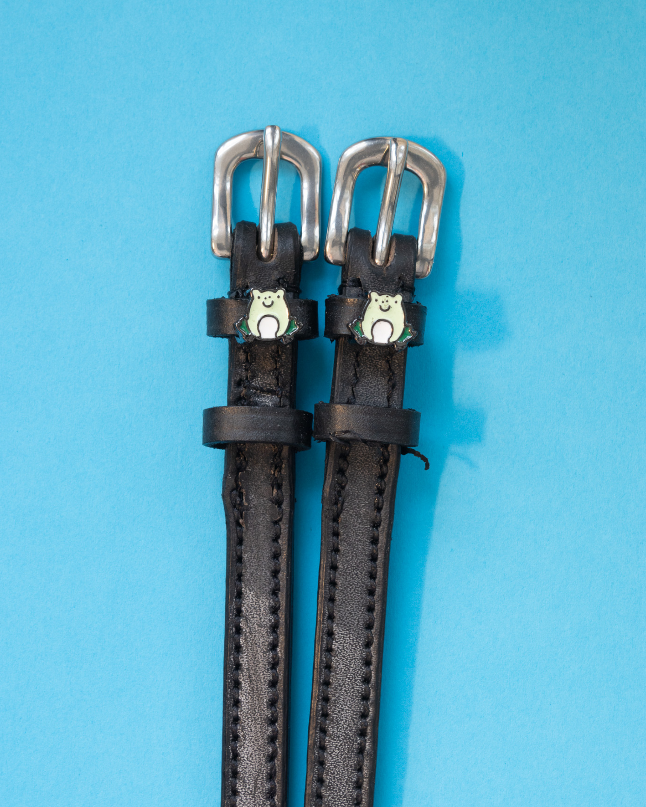 Black leather ManeJane frog spur straps with silver buckles on a light blue background