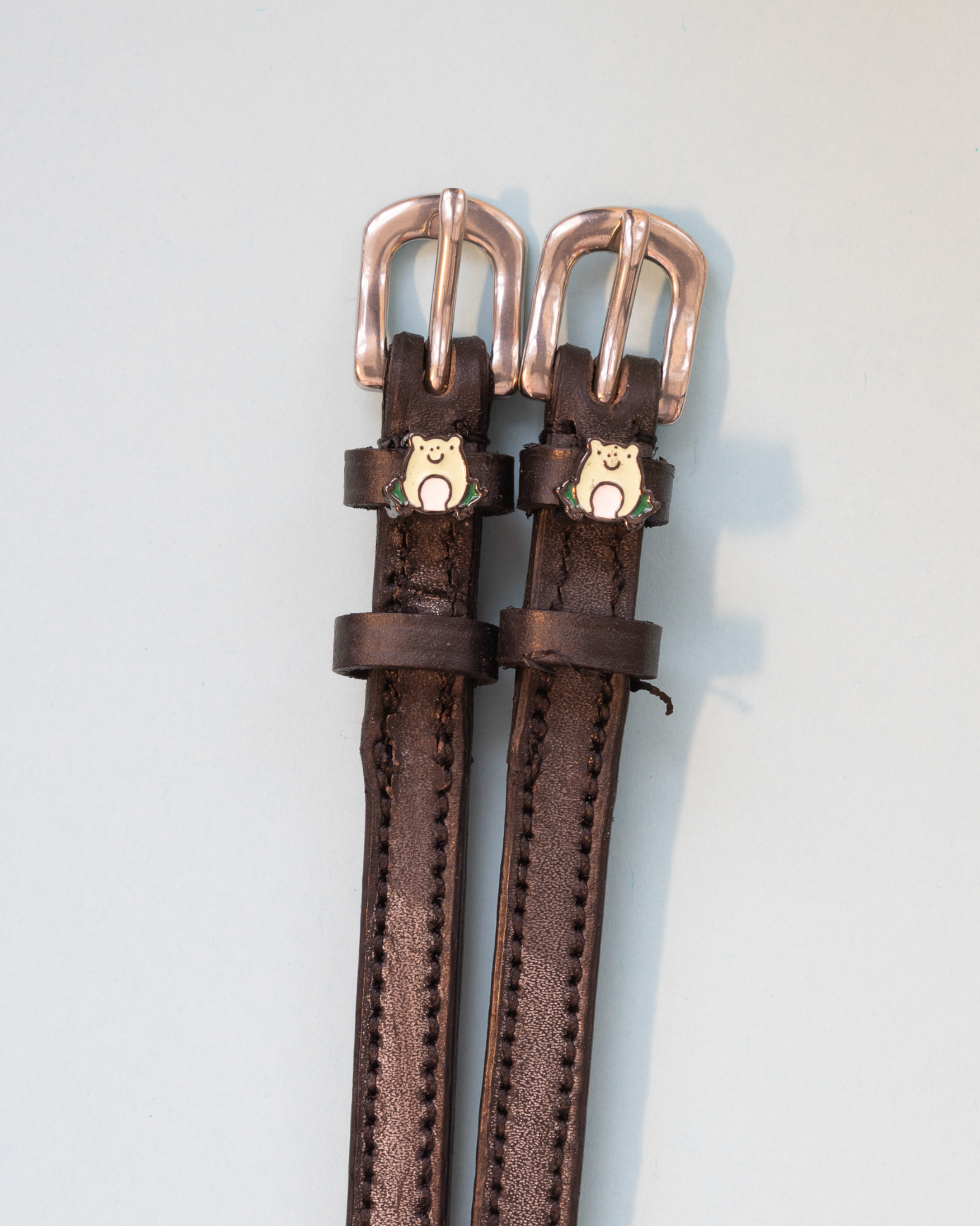 Brown leather ManeJane frog spur straps with rose-gold buckles on a grey background