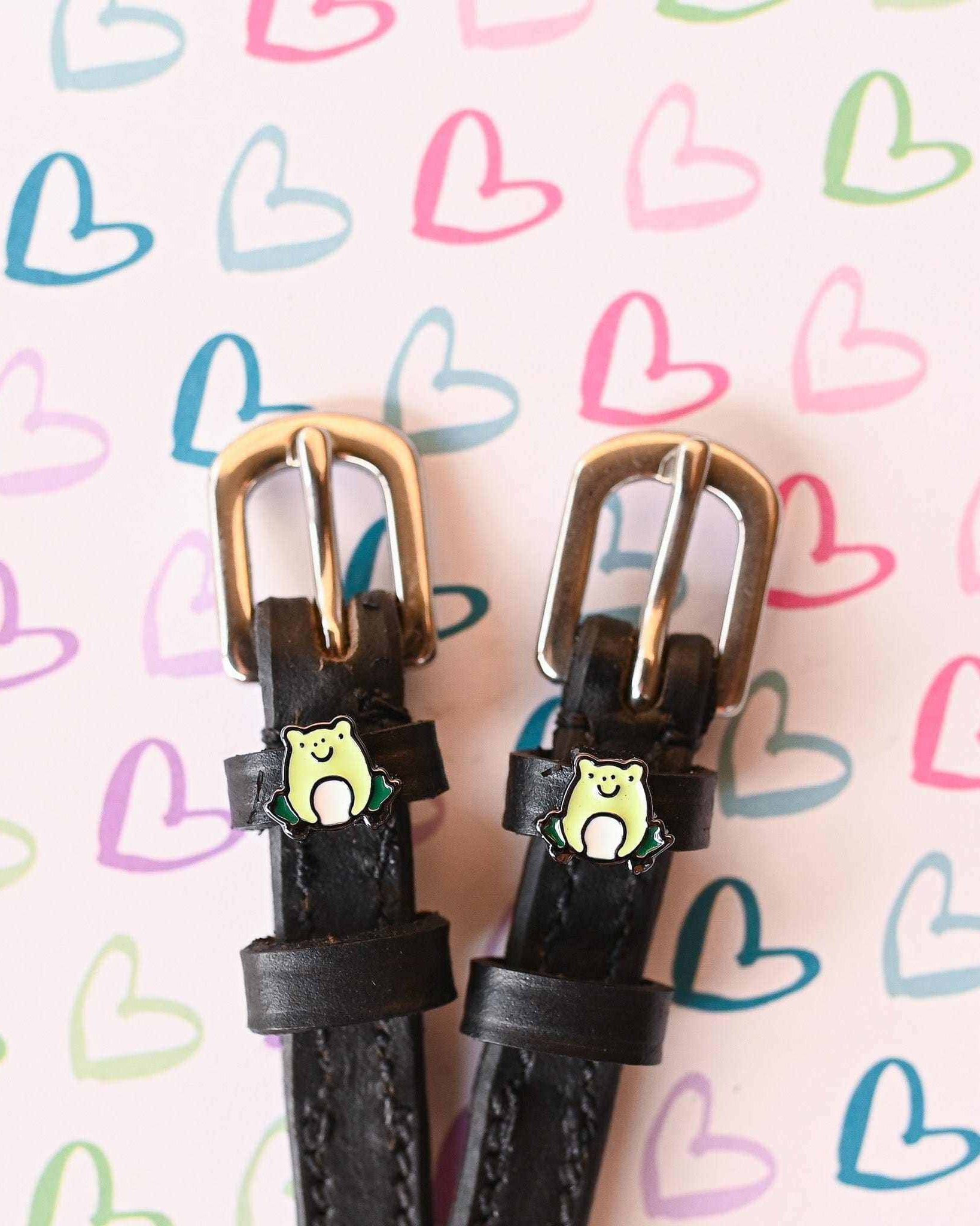 Black leather frog spur straps with brass buckles on a pastel heart-patterned background