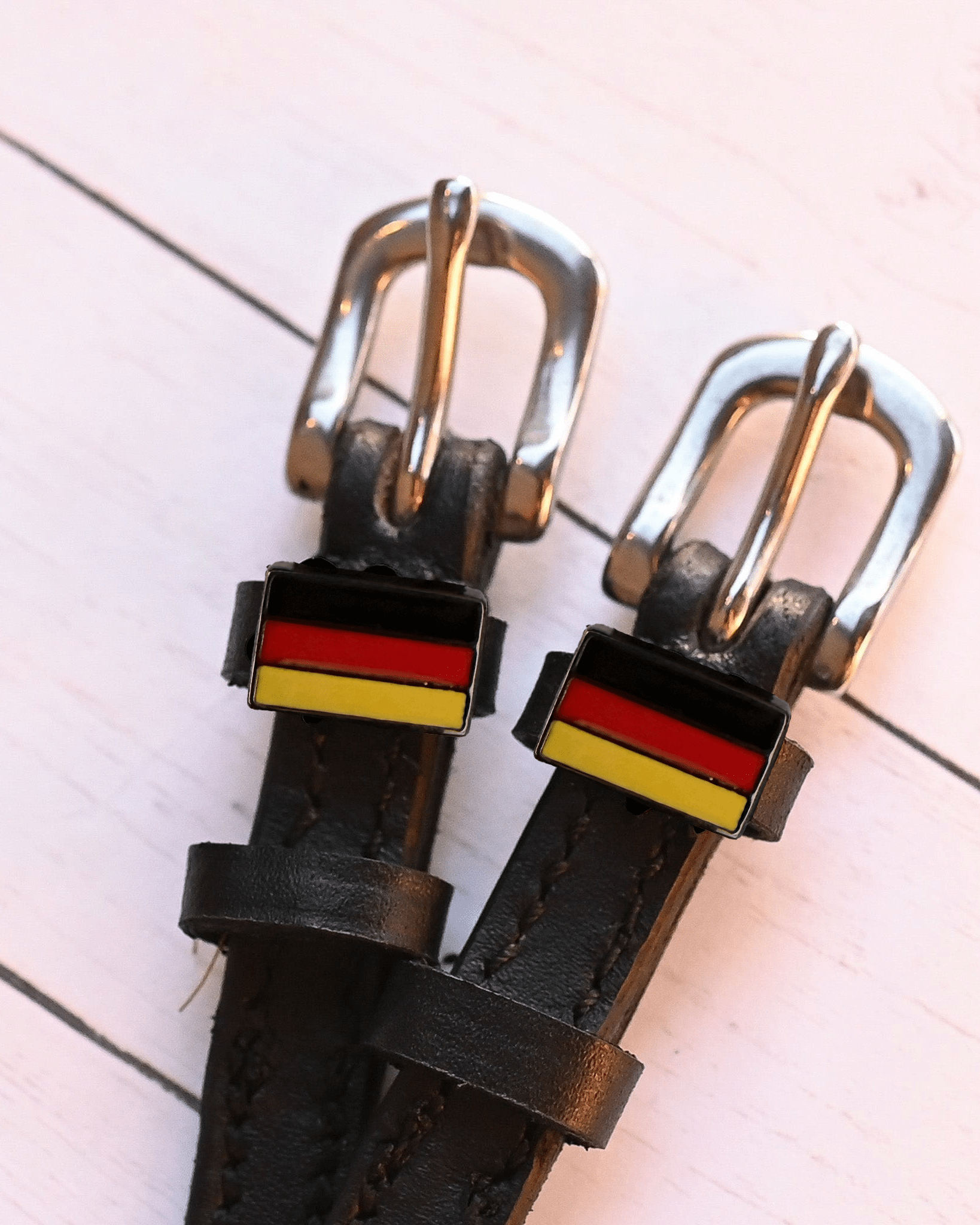 German flag charm spur straps in black leather on a pale pink wood-plank background
