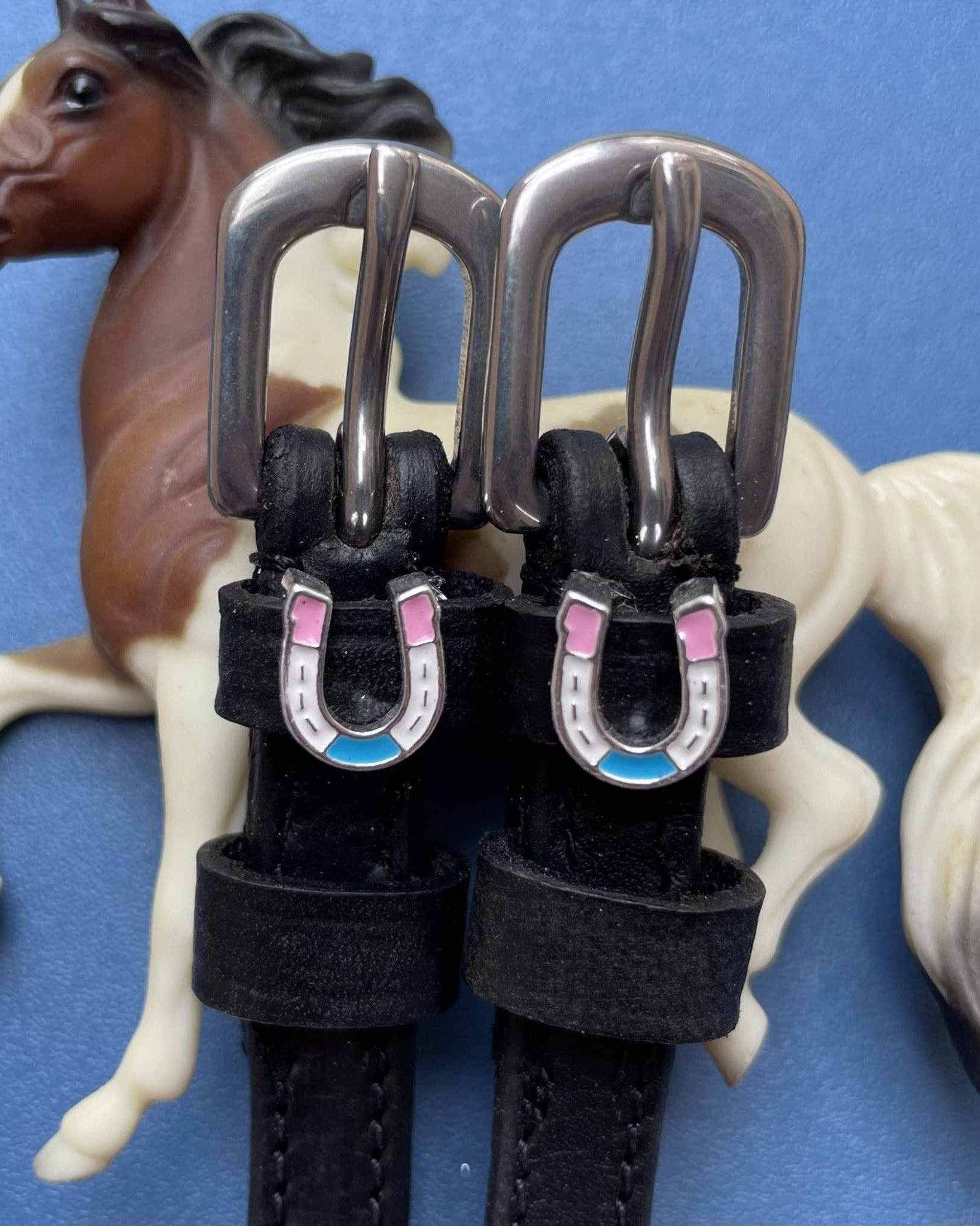 Pastel horseshoe charm spur straps styled with a toy horse figurine on blue