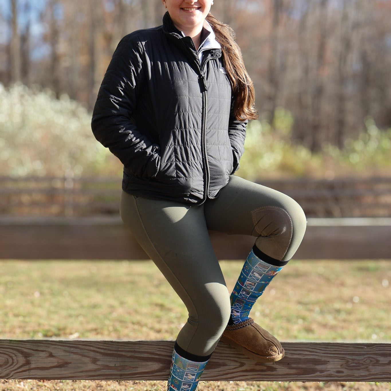 Woman sitting on wooden fence wearing Limited Edition Pony Island Holiday Barn Socks with festive pony design