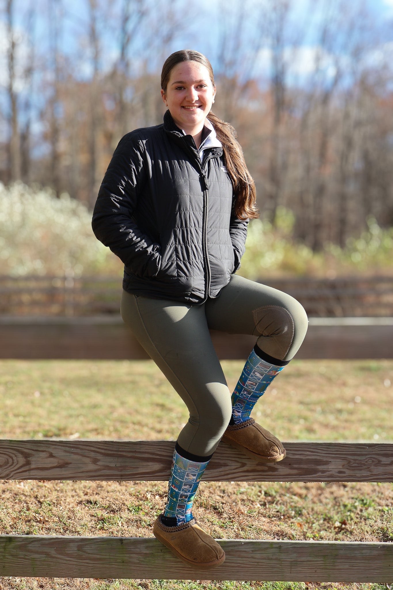 Woman sitting on wooden fence wearing Limited Edition Pony Island Holiday Barn Socks with festive pony design